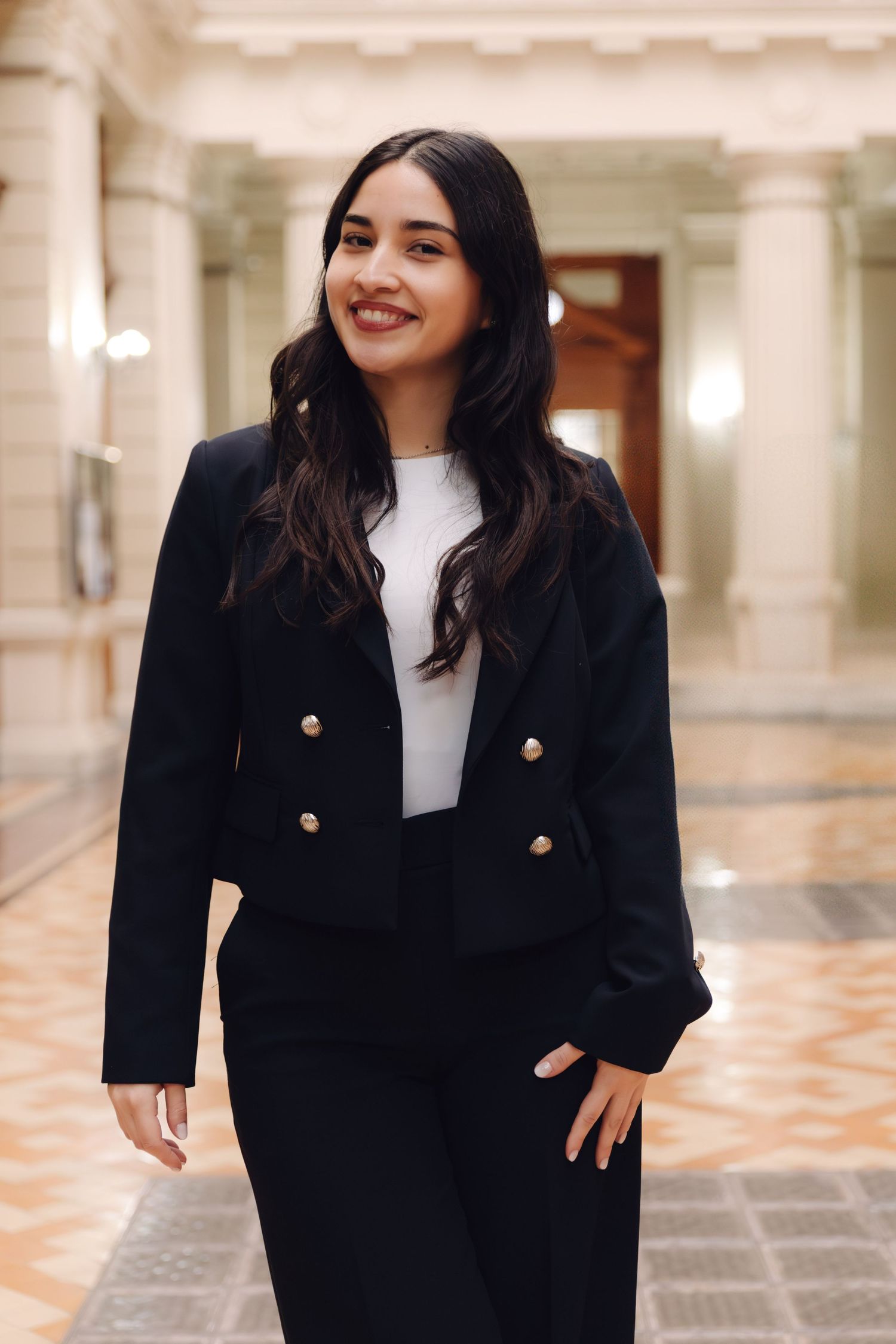 Young woman in black double-breasted blazer smiling confidently while walking through an elegant interior hallway.