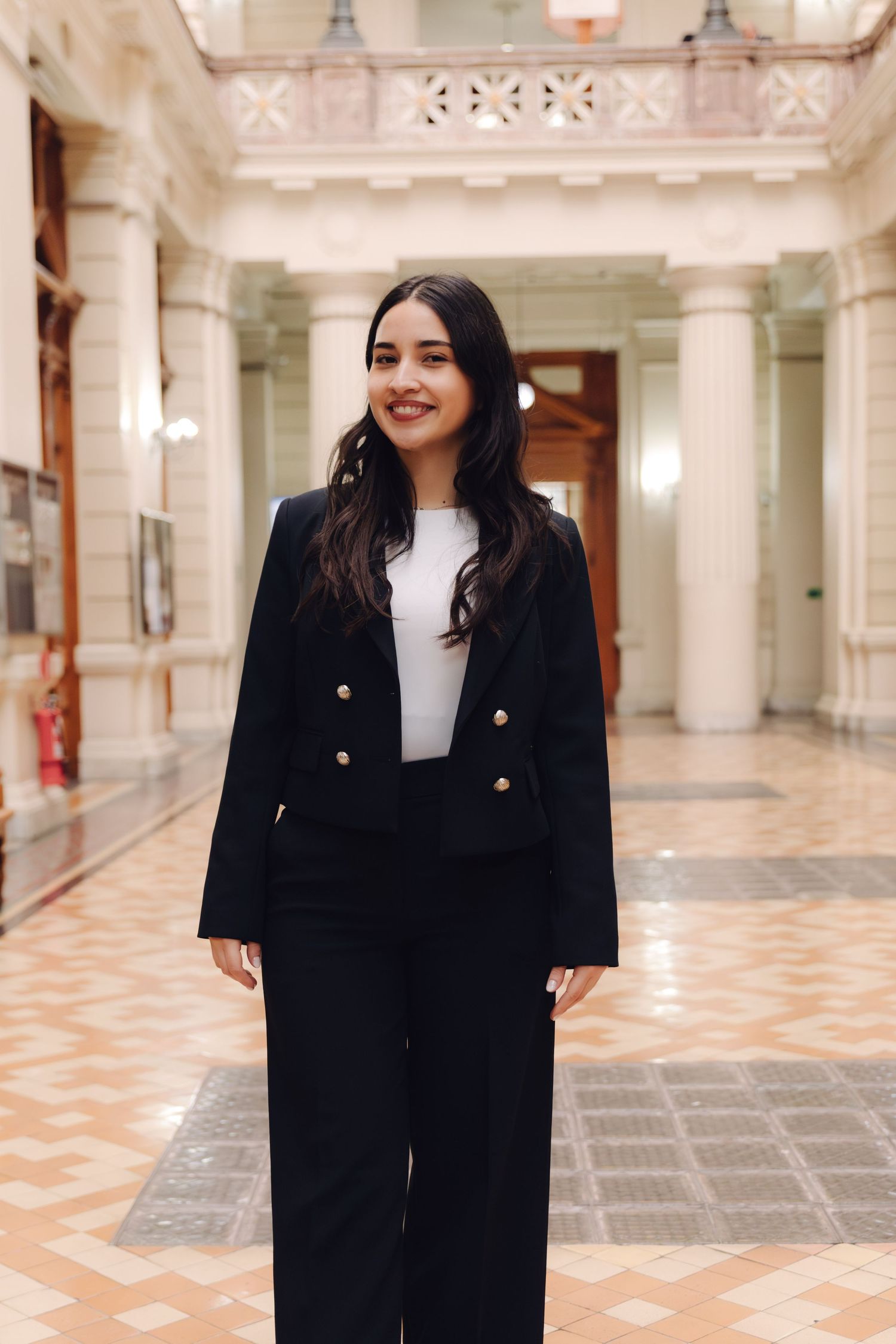 Young woman in black blazer and wide-leg pants smiling in a grand columned interior hallway.