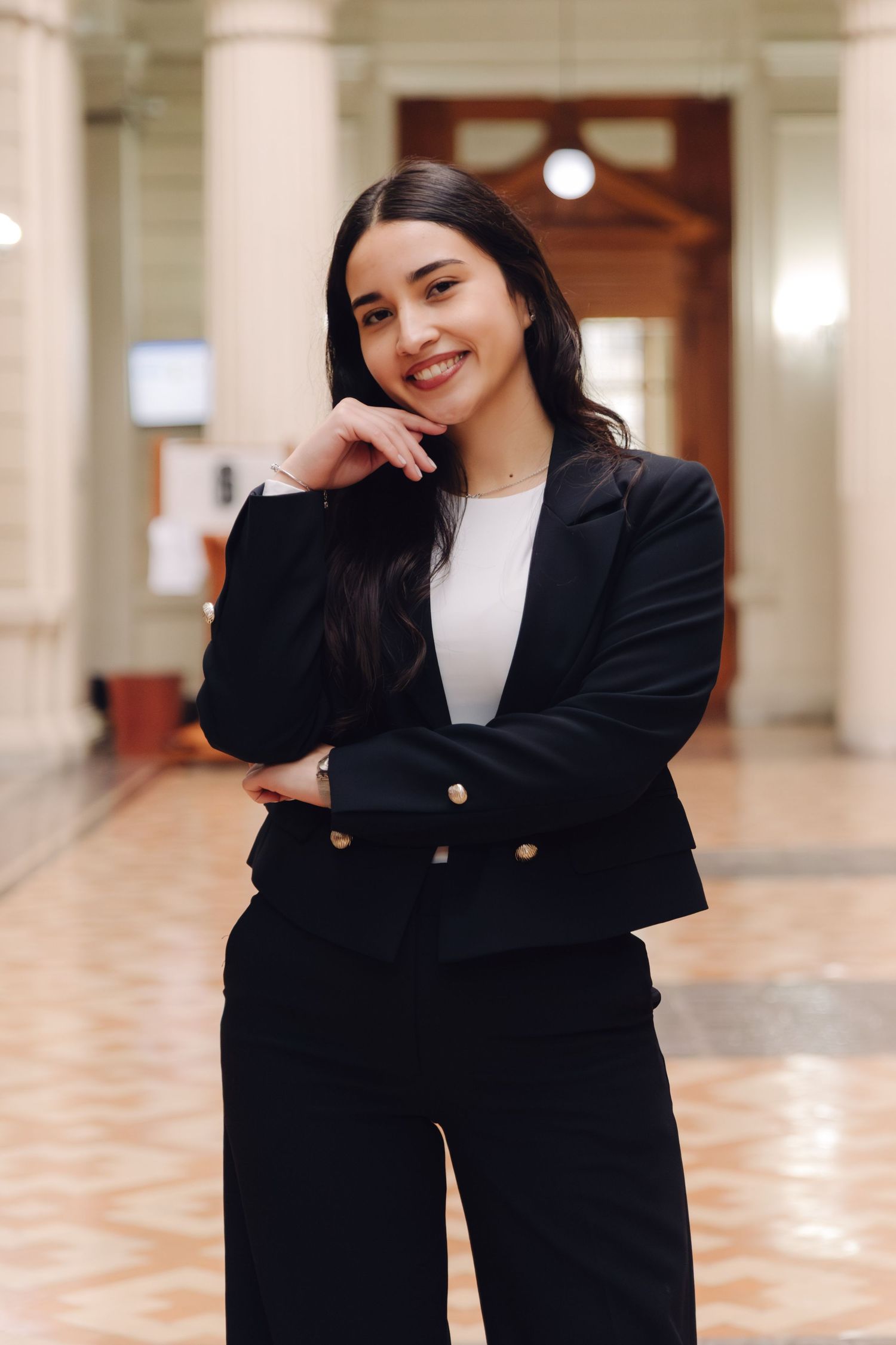 Professional woman in black suit smiling with hand on chin in ornate building interior.