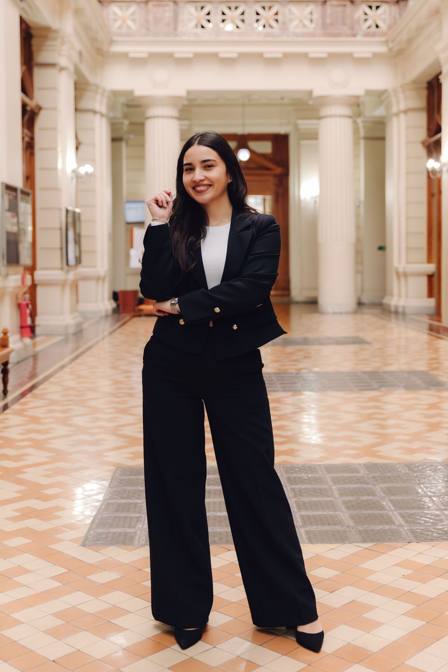 Young woman in black blazer and wide-leg pants smiling in elegant hall with columns.