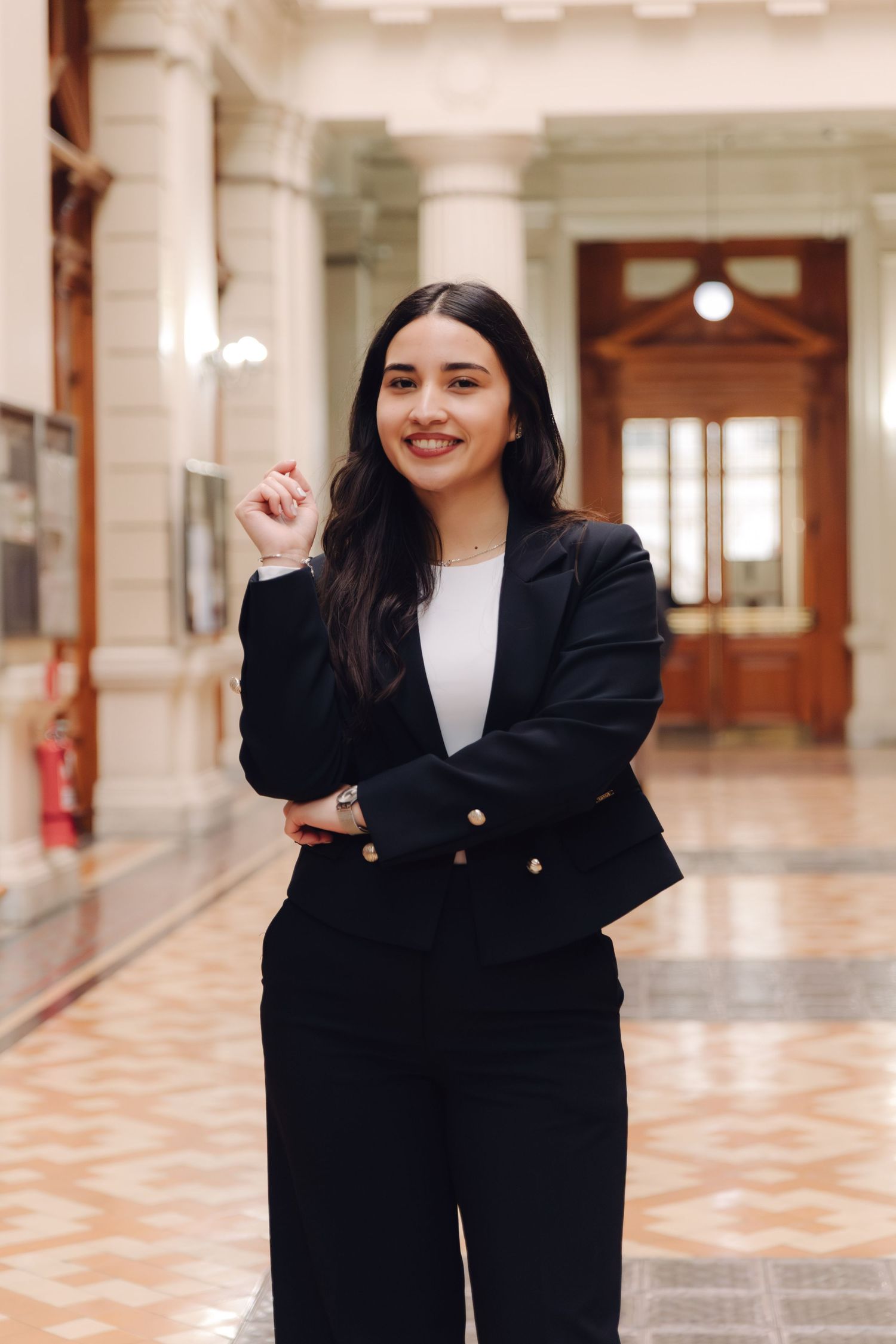 Smiling young woman in black blazer posing confidently in grand architectural hallway.