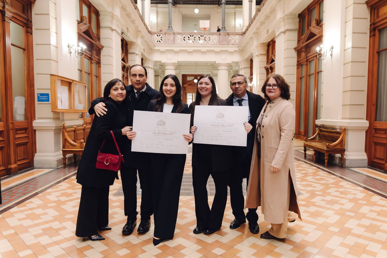 Group of six people posing with diplomas in elegant corridor celebrating graduation.