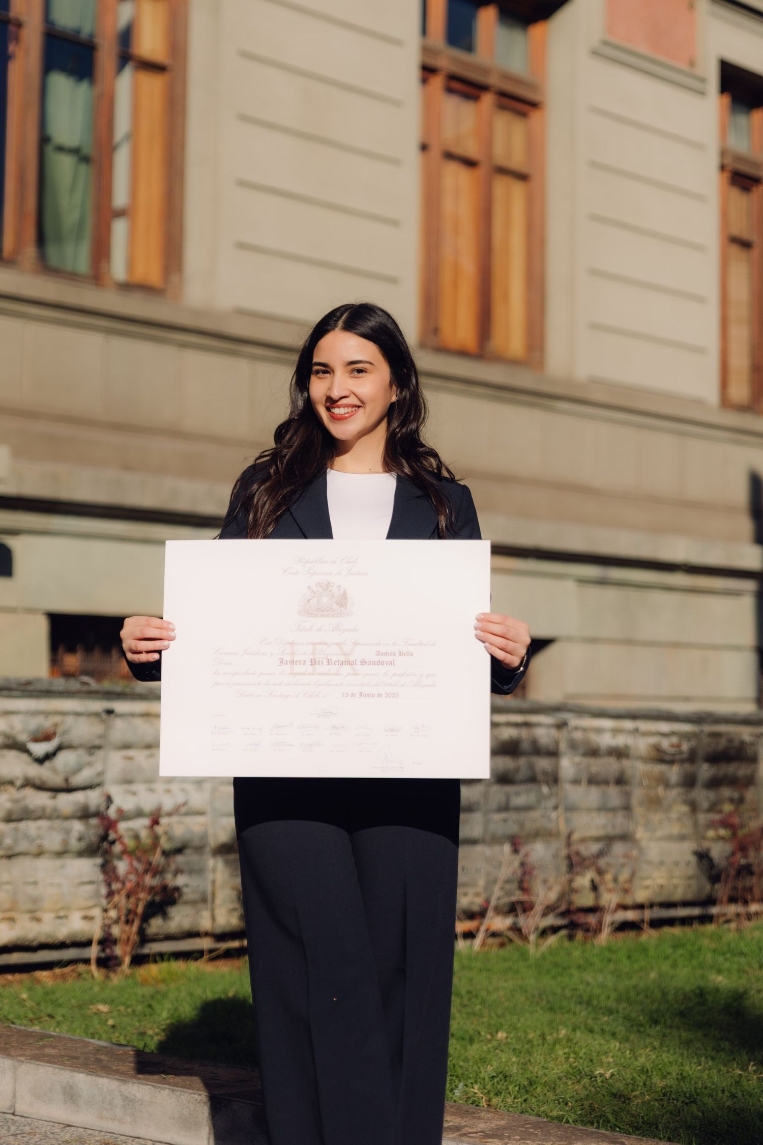 Smiling graduate in black suit holding diploma certificate outdoors near historic building.