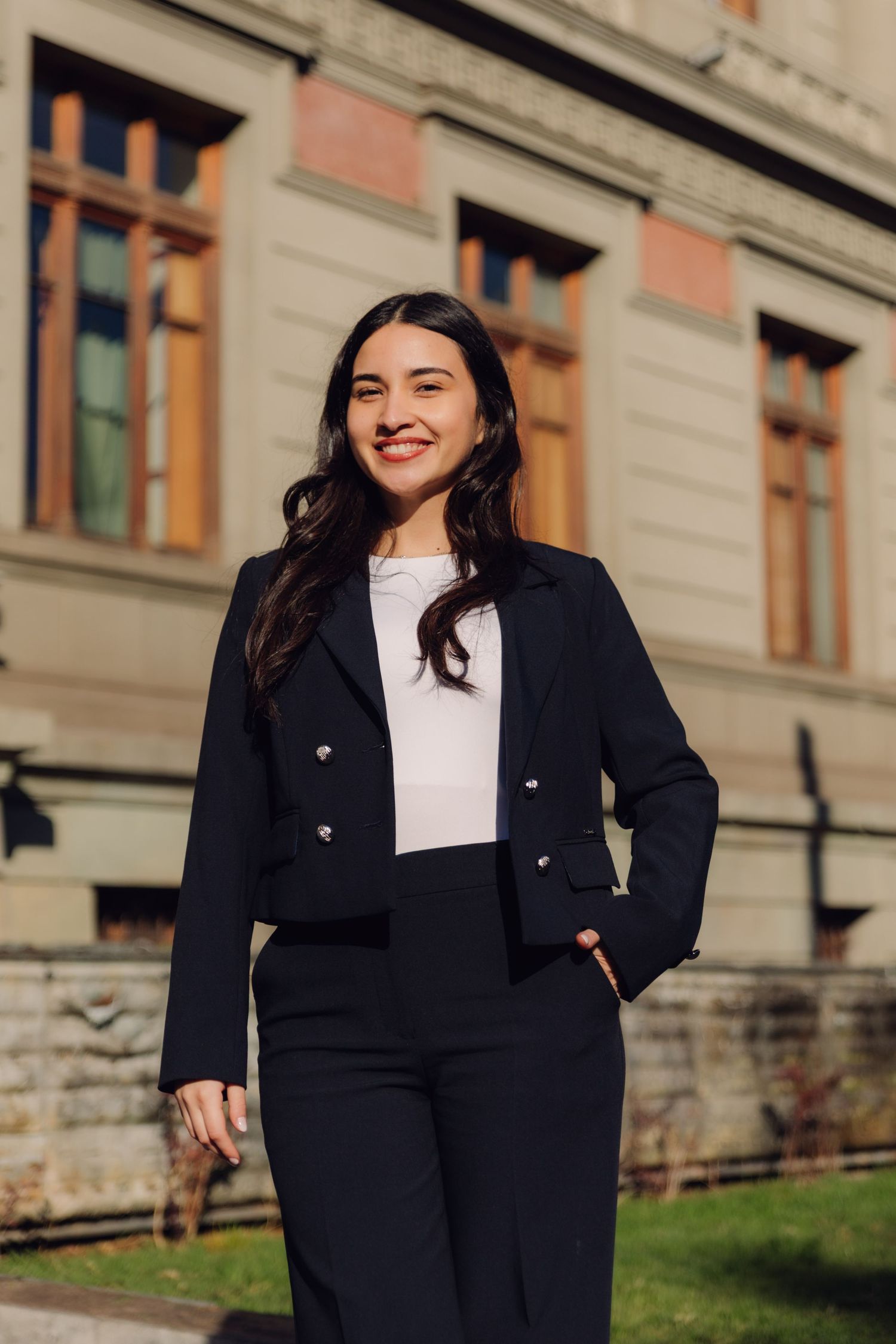Young professional woman in black blazer smiling outdoors near classic stone building.