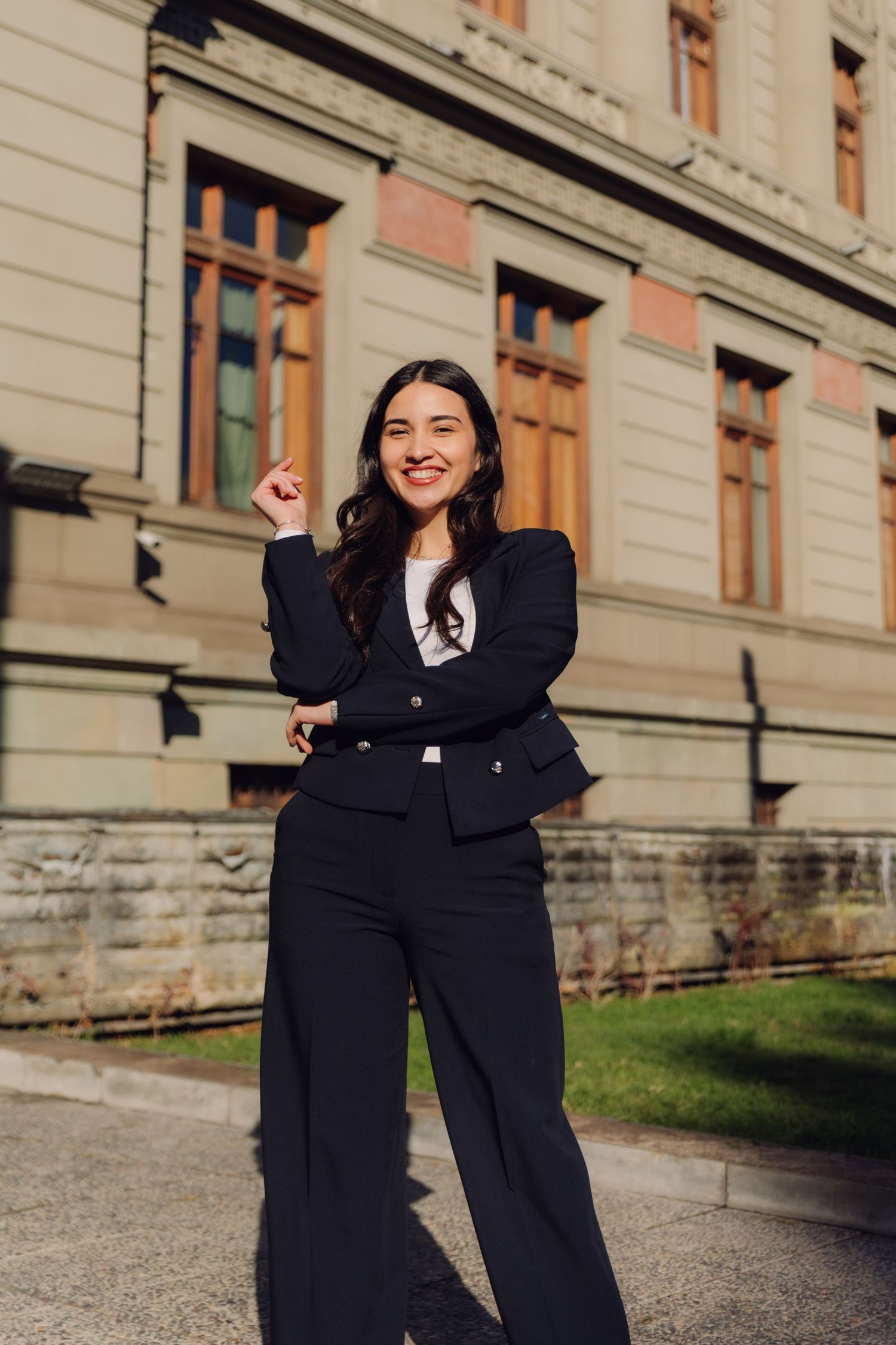 Cheerful woman in black pantsuit posing with raised fist outside historic building.