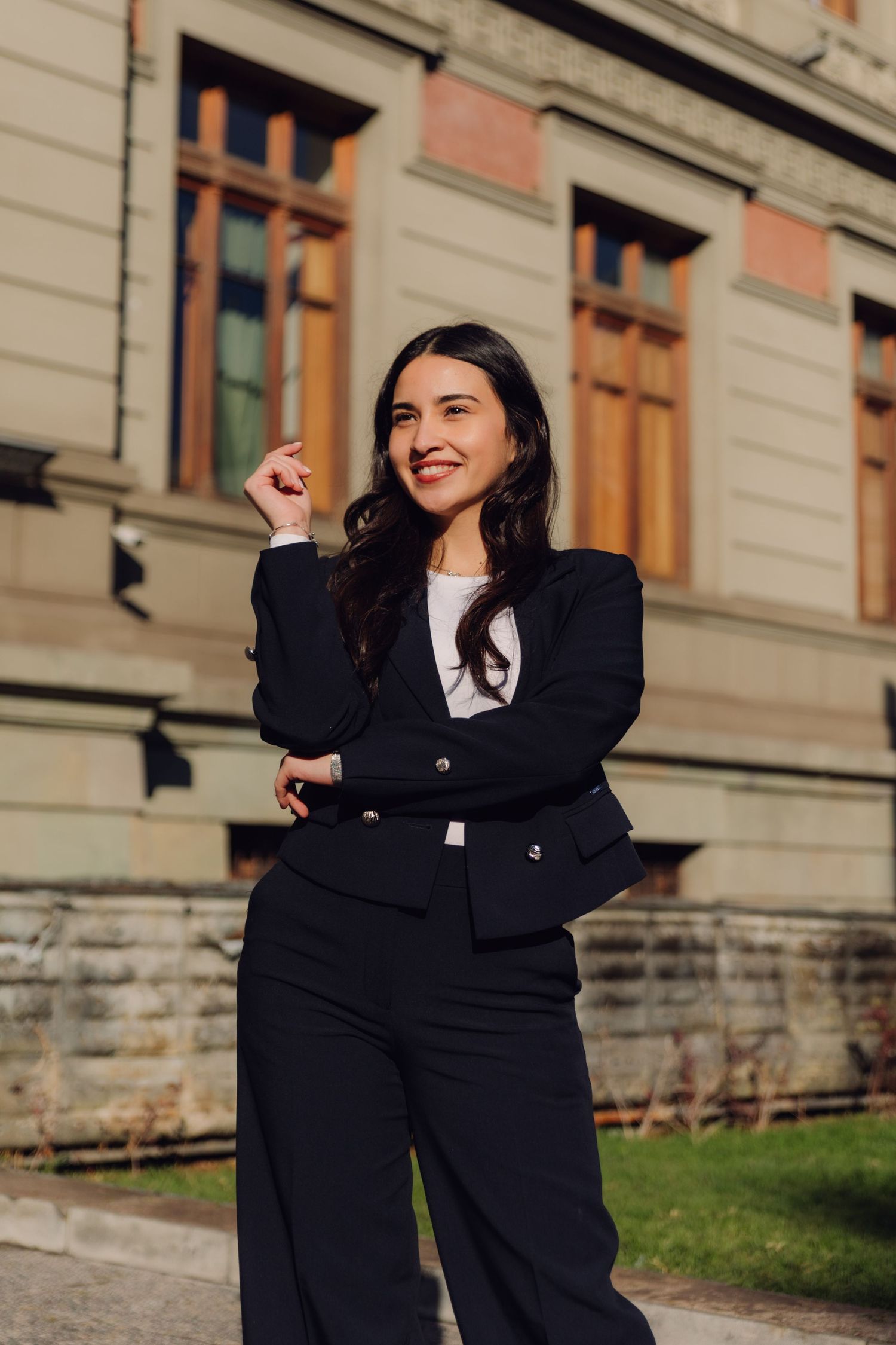 Confident young woman in black suit smiling outdoors beside classic stone architecture.