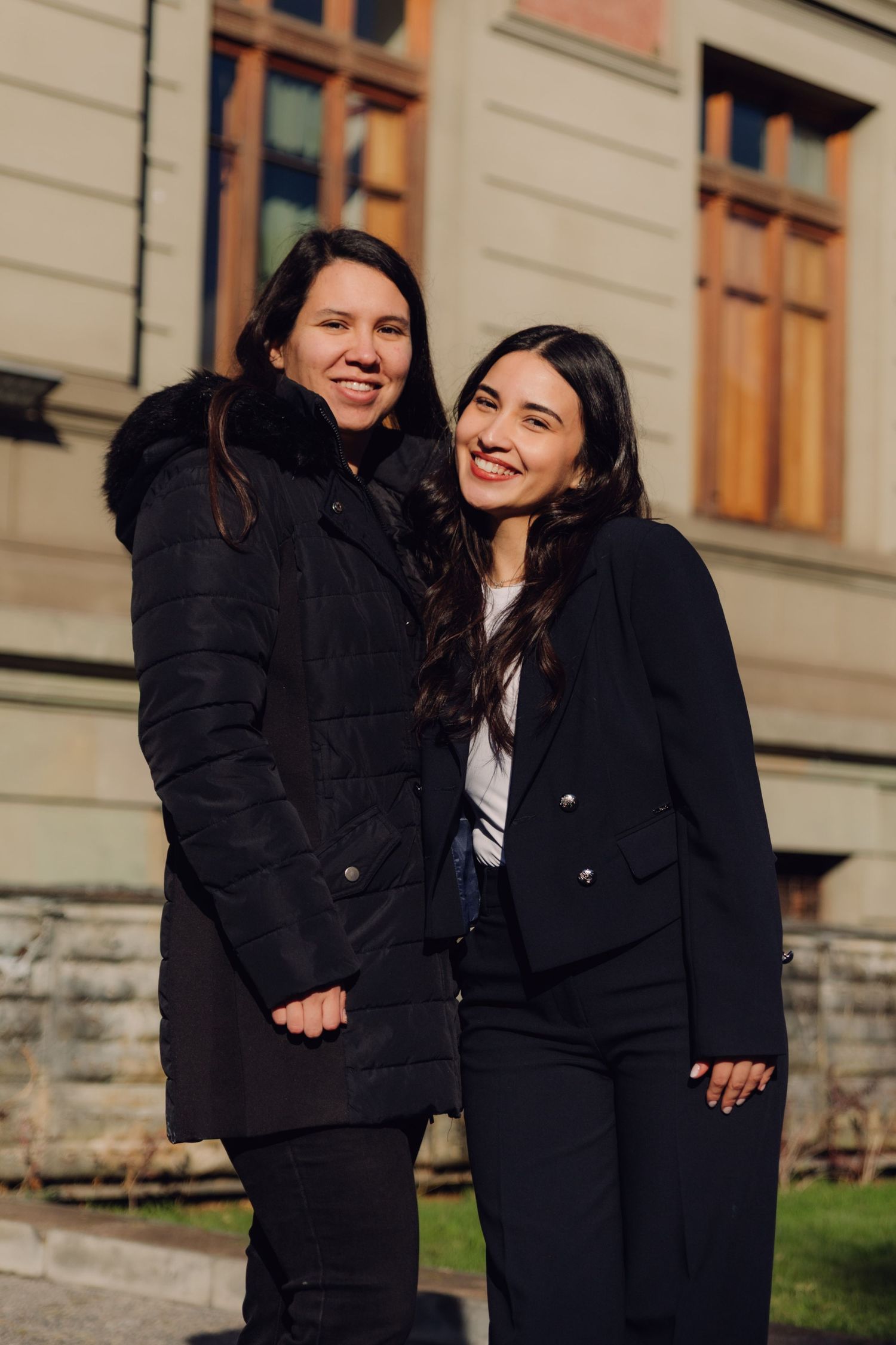 Two smiling women in black coats posing together outside historic stone building.