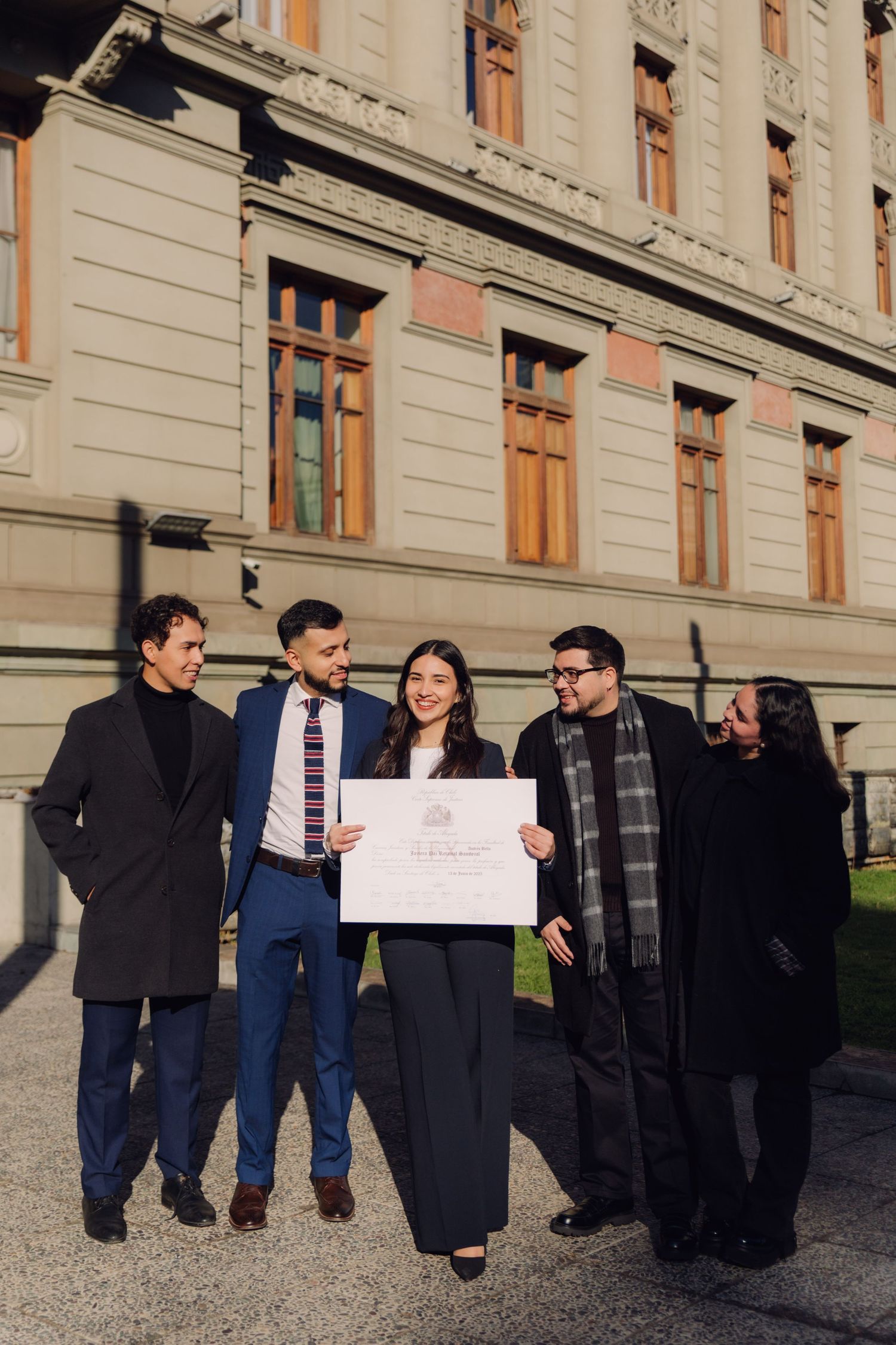 Graduate holding diploma surrounded by family celebrating outside elegant stone building.