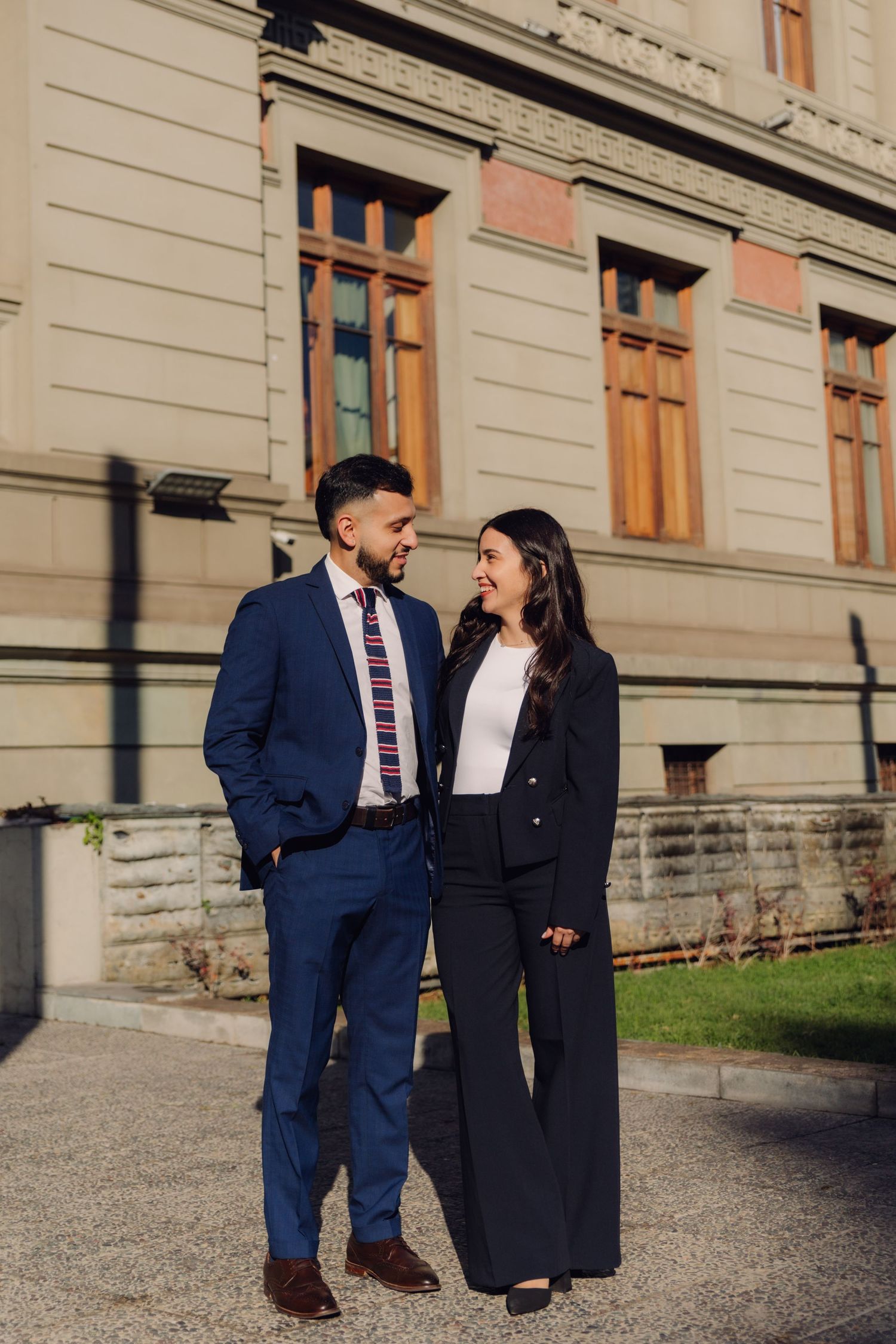 A man in a blue suit and woman in black blazer smiling at each other outside a classic building.