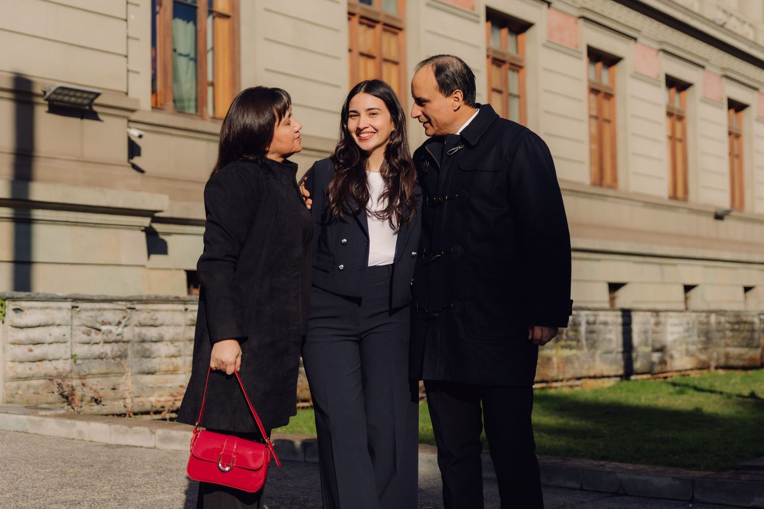 Three people smiling and conversing outdoors near a classic stone building on a sunny day.