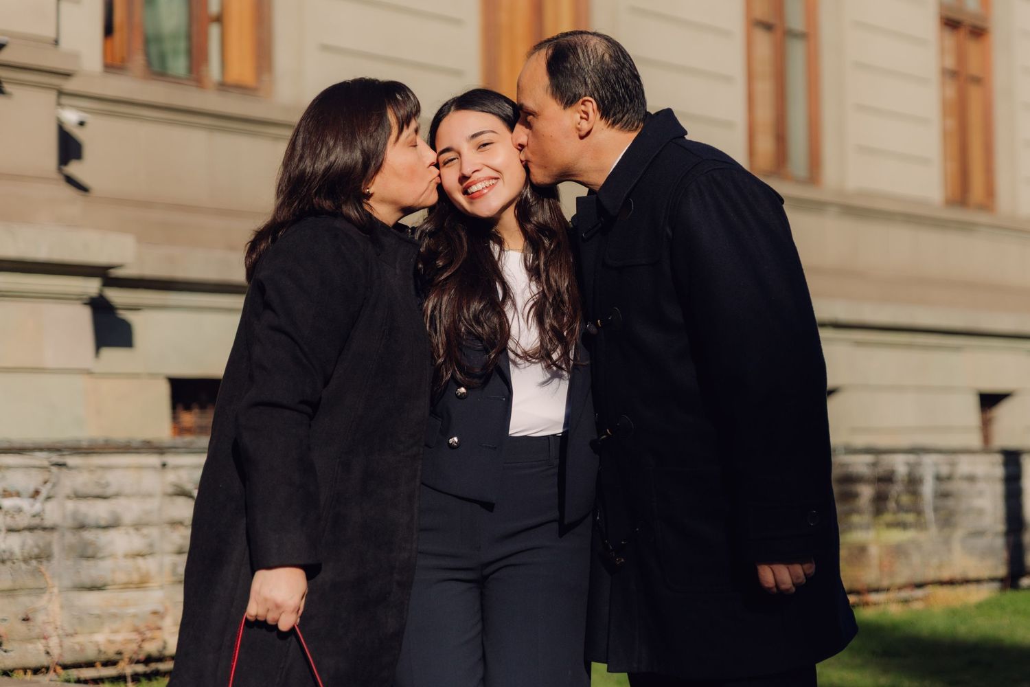 Two older adults kissing a young woman on the cheeks outdoors near a historic building.