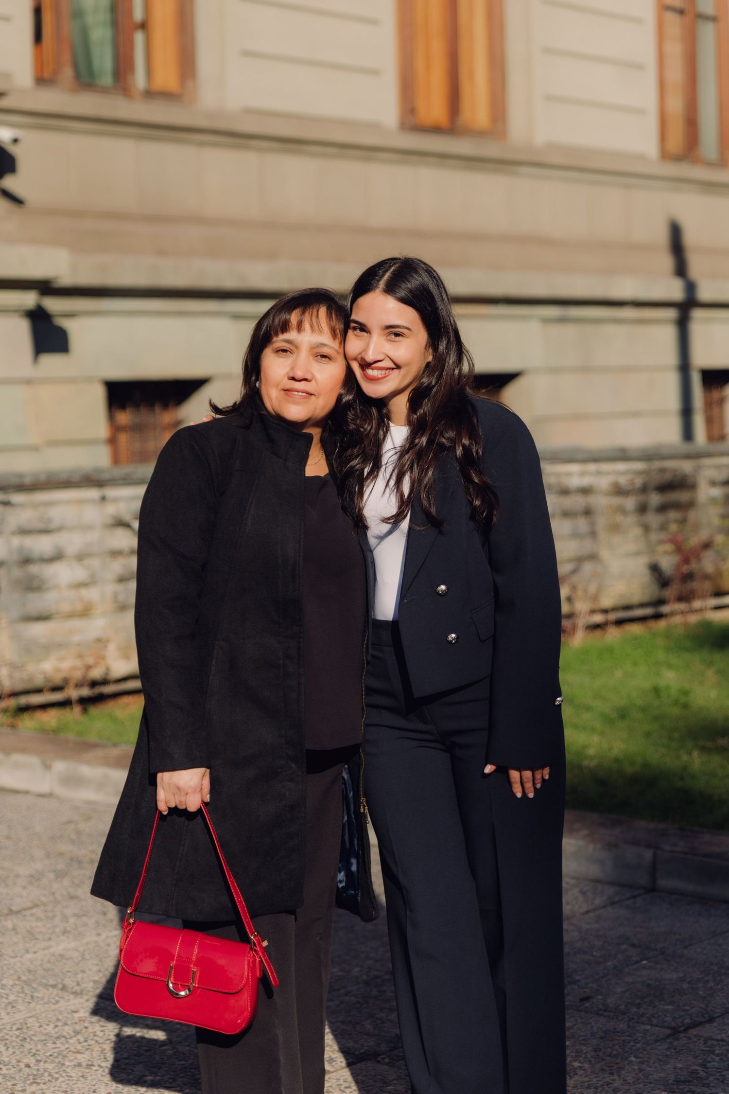 Two women in black outfits smiling together outside a classic building on a sunny day.