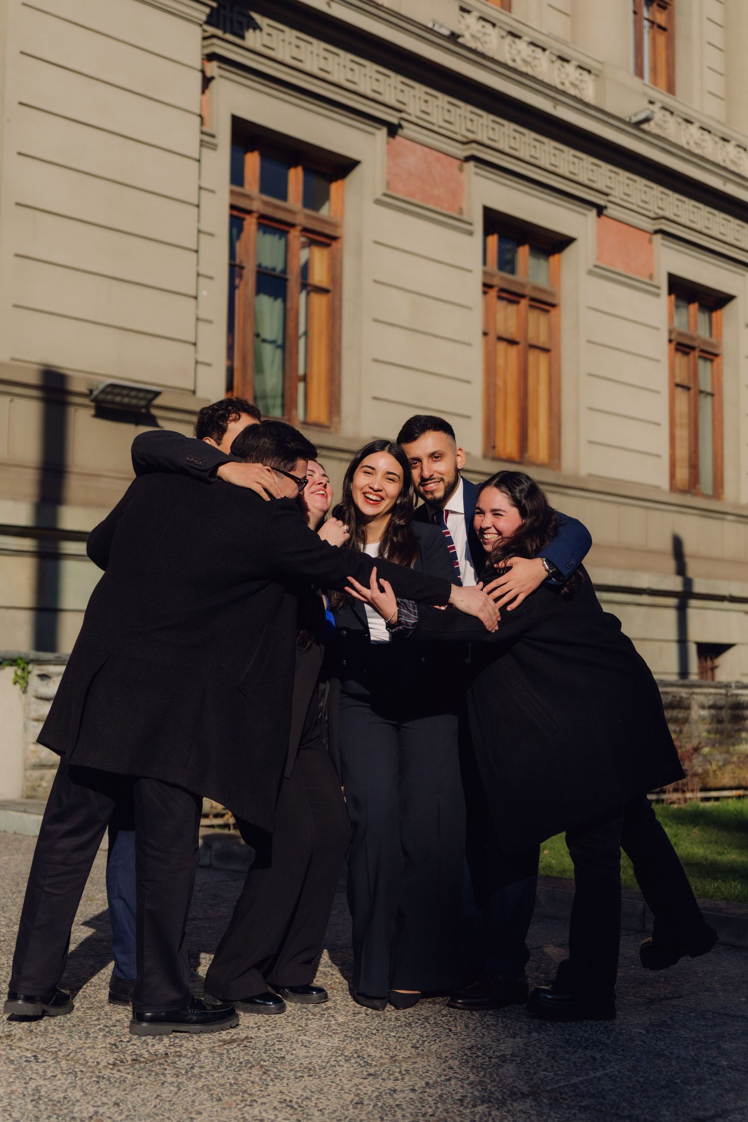 Five people laughing and embracing in a group hug outside a classic European building.