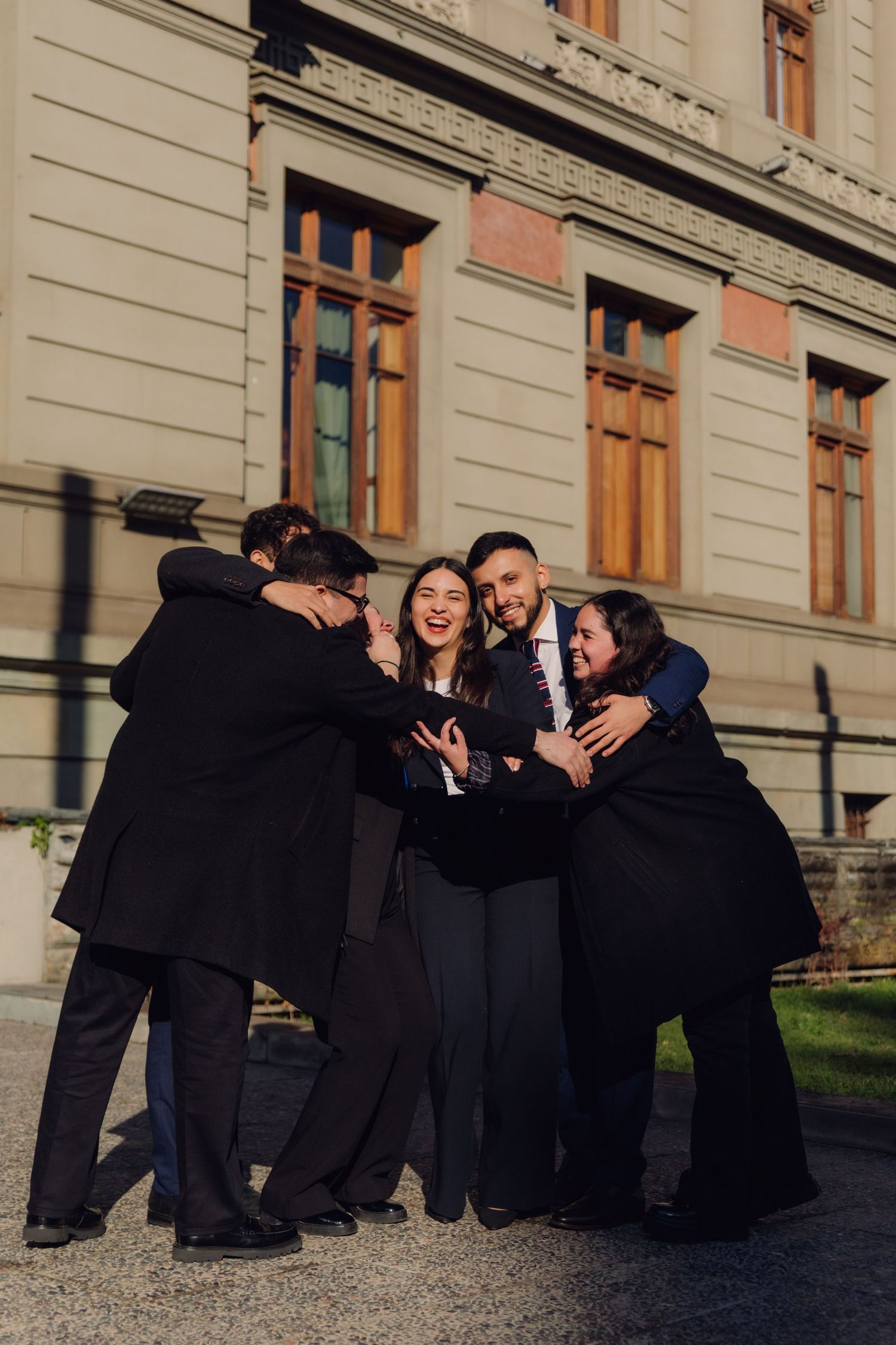 A joyful group of five people sharing a group hug outside a historic stone building.