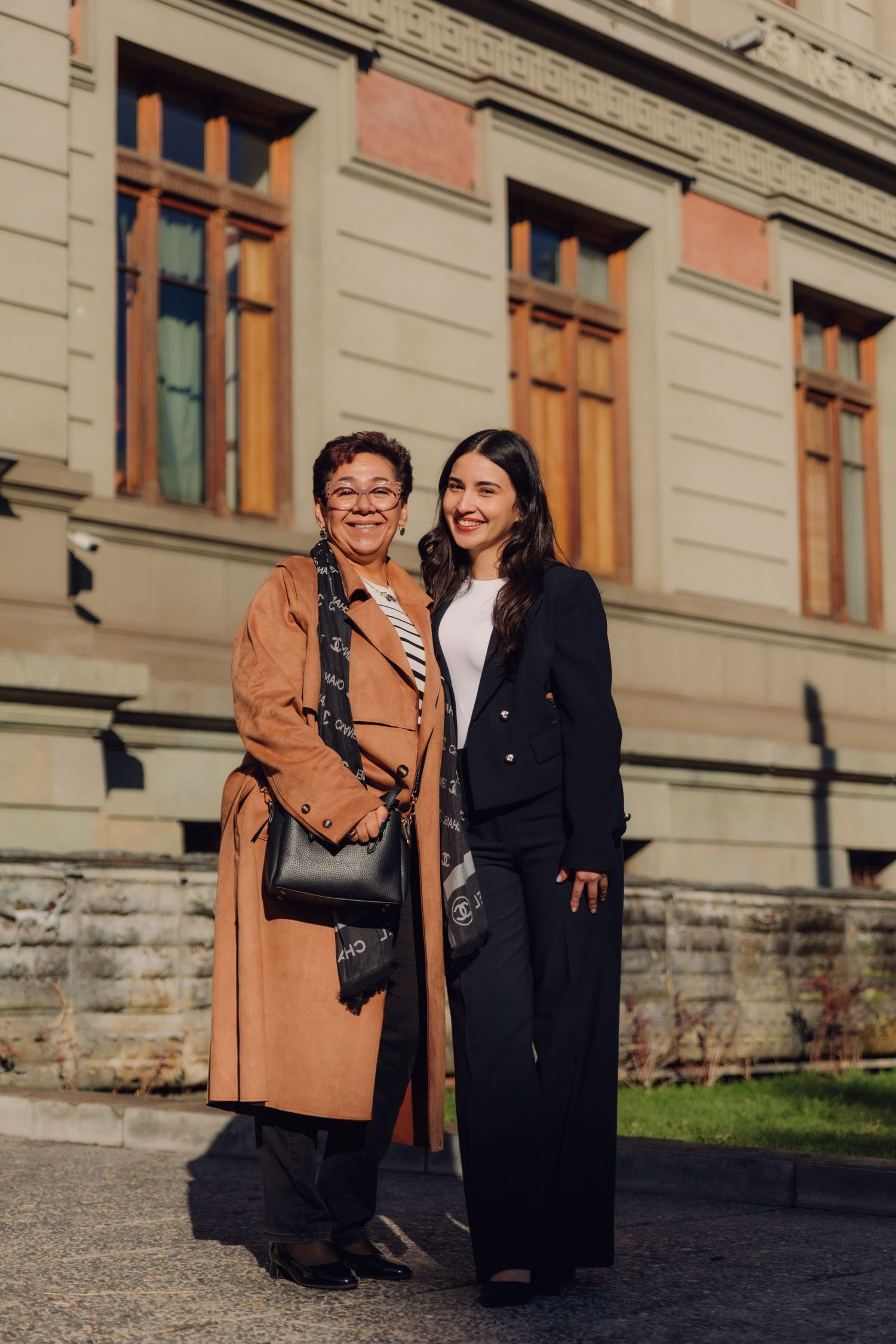 Two women in elegant coats smiling together outside a sunlit historic building.