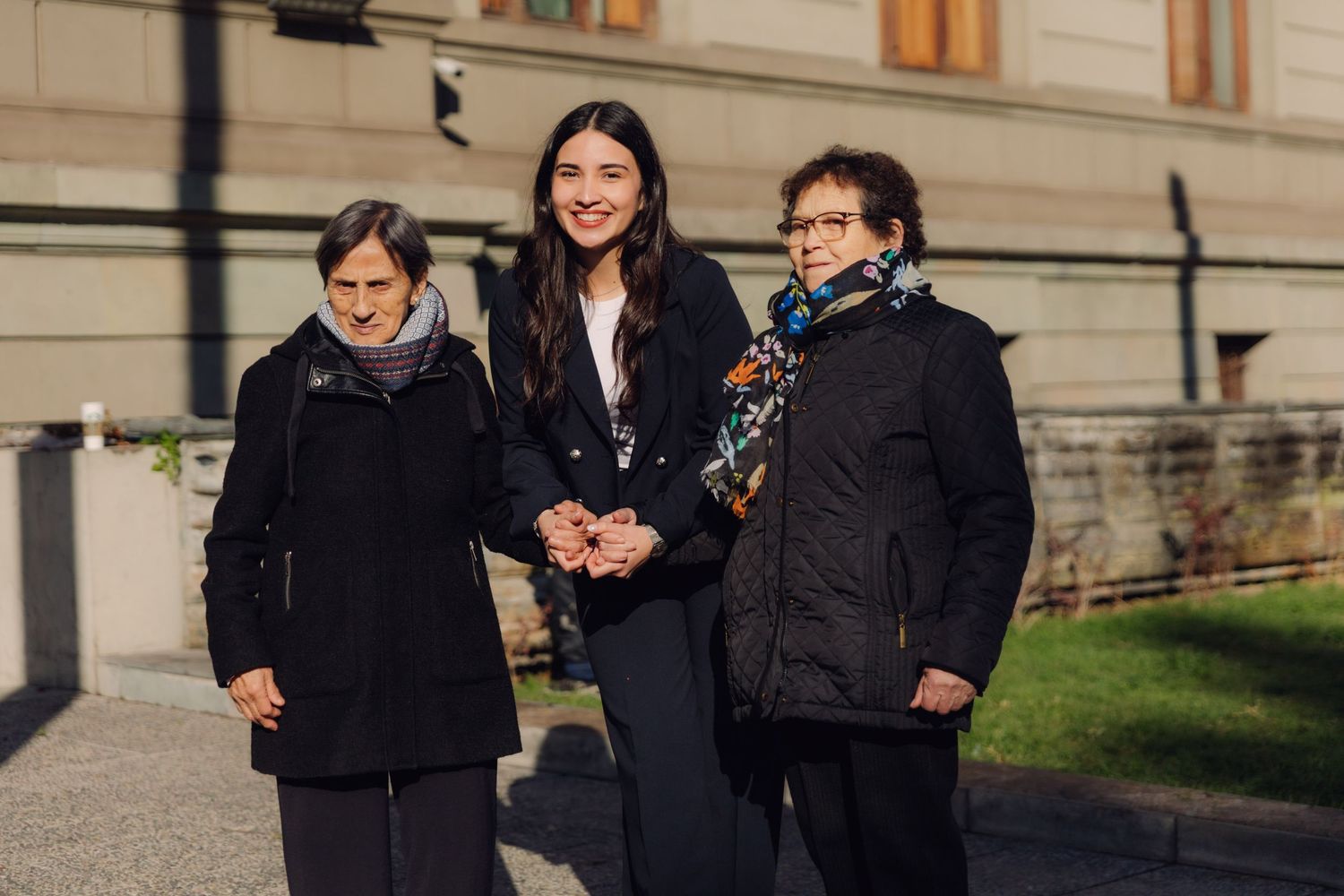 A young woman in a navy blazer standing between two older women outside a classic building.