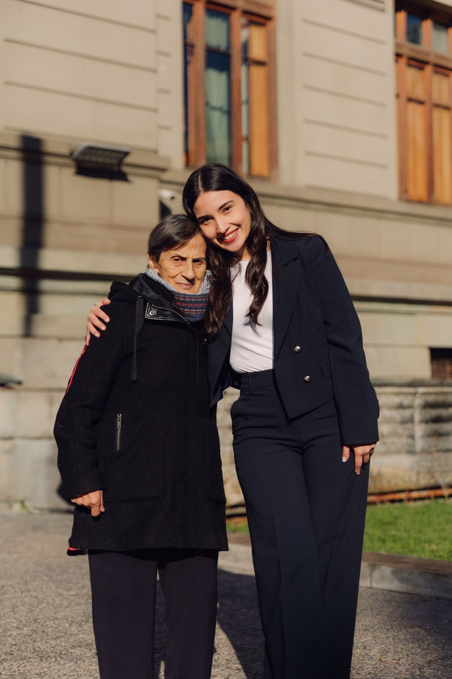 A young woman in a navy blazer embracing an elderly woman outside a classic stone building.