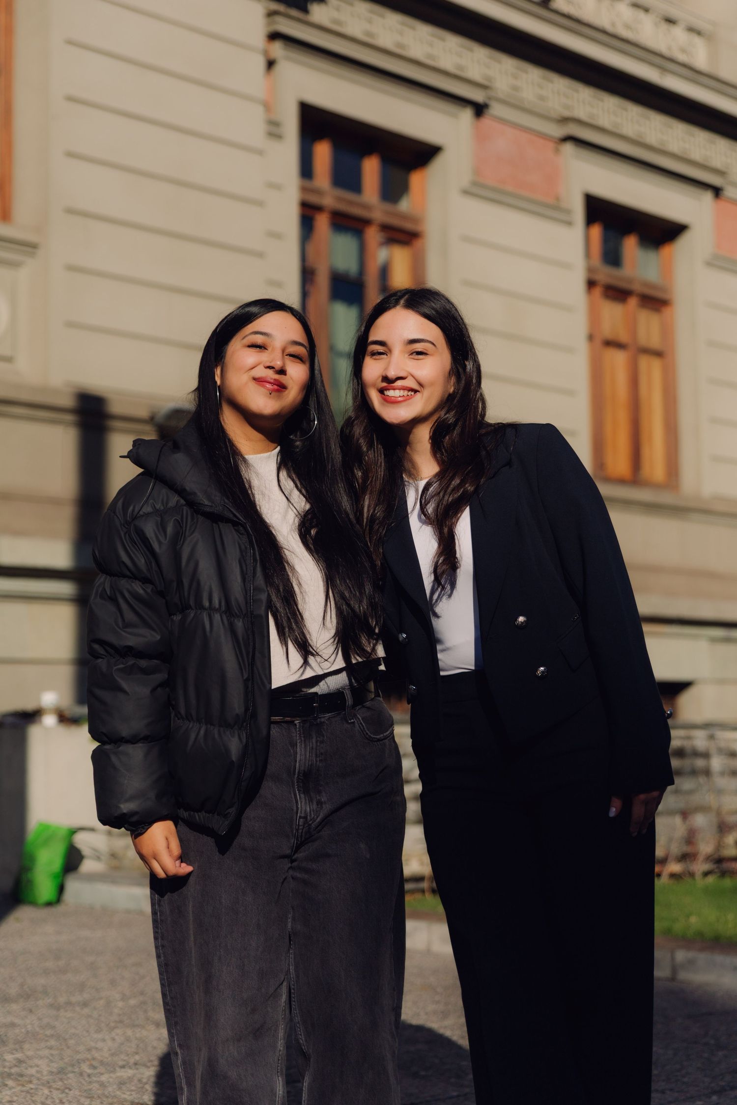 Two young women in black outfits smiling outdoors near a classic building.