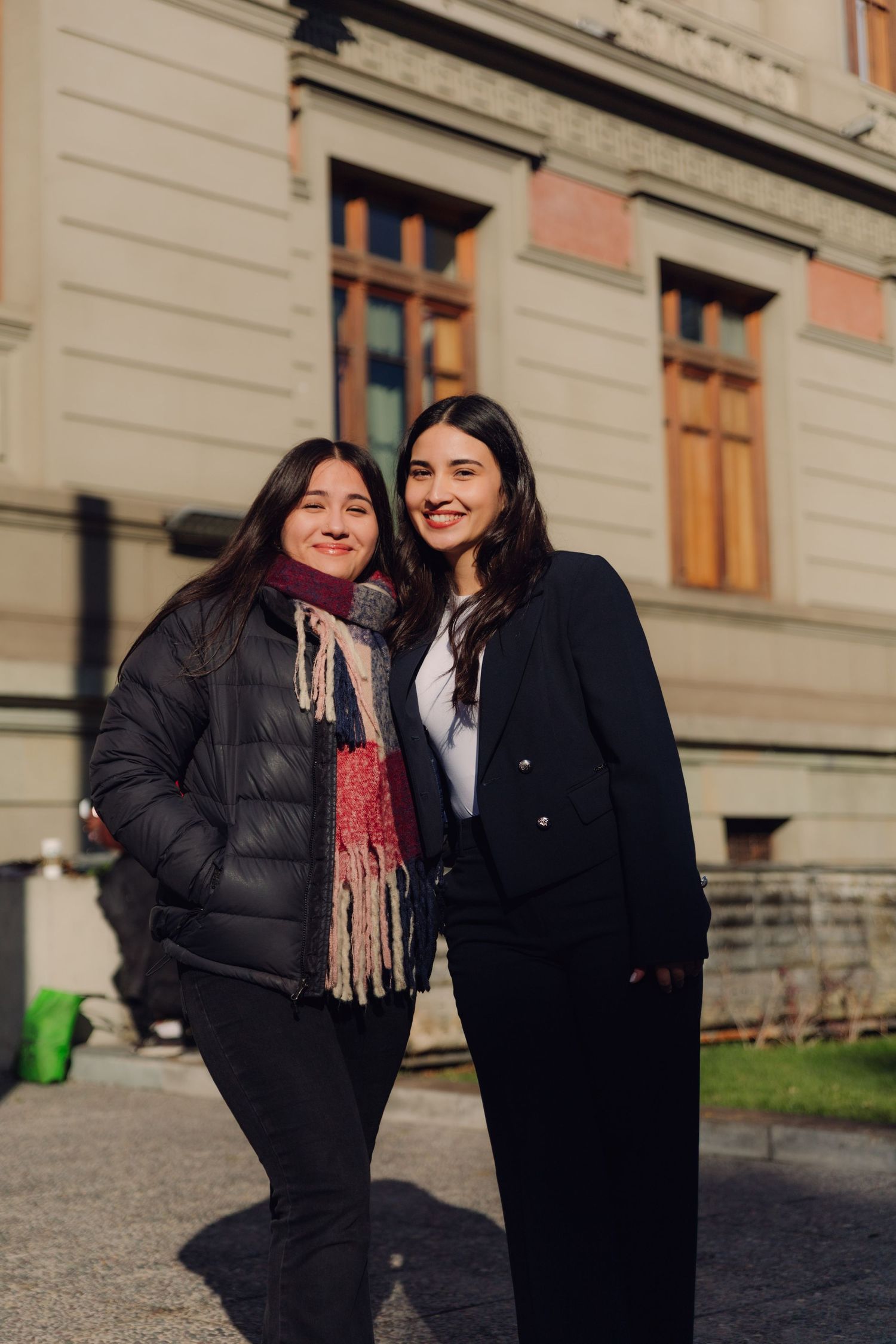 Two smiling women posing together outside a historic building on a sunny day.
