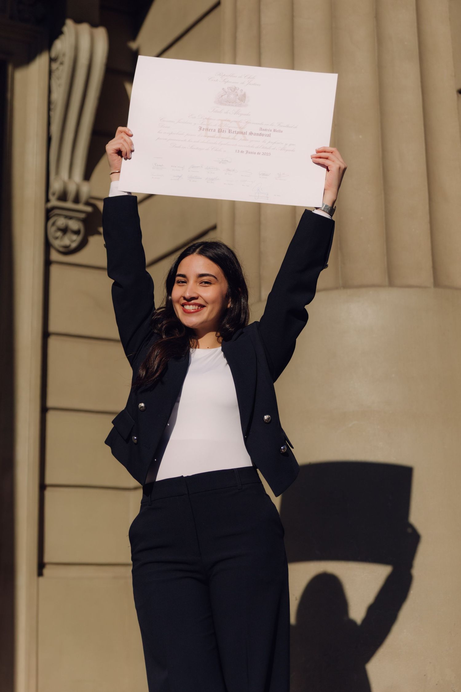 Young woman in black suit celebrating by holding diploma above her head outdoors.