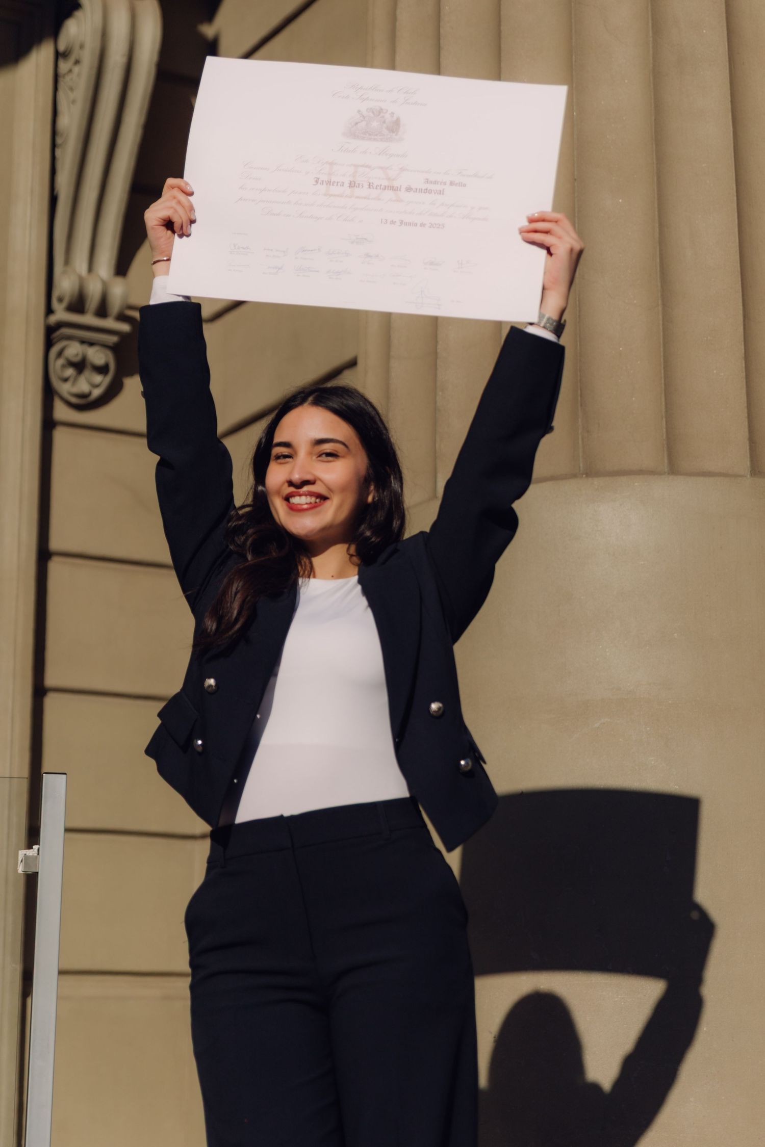 Smiling graduate in black blazer proudly raising her diploma outside a classic building.