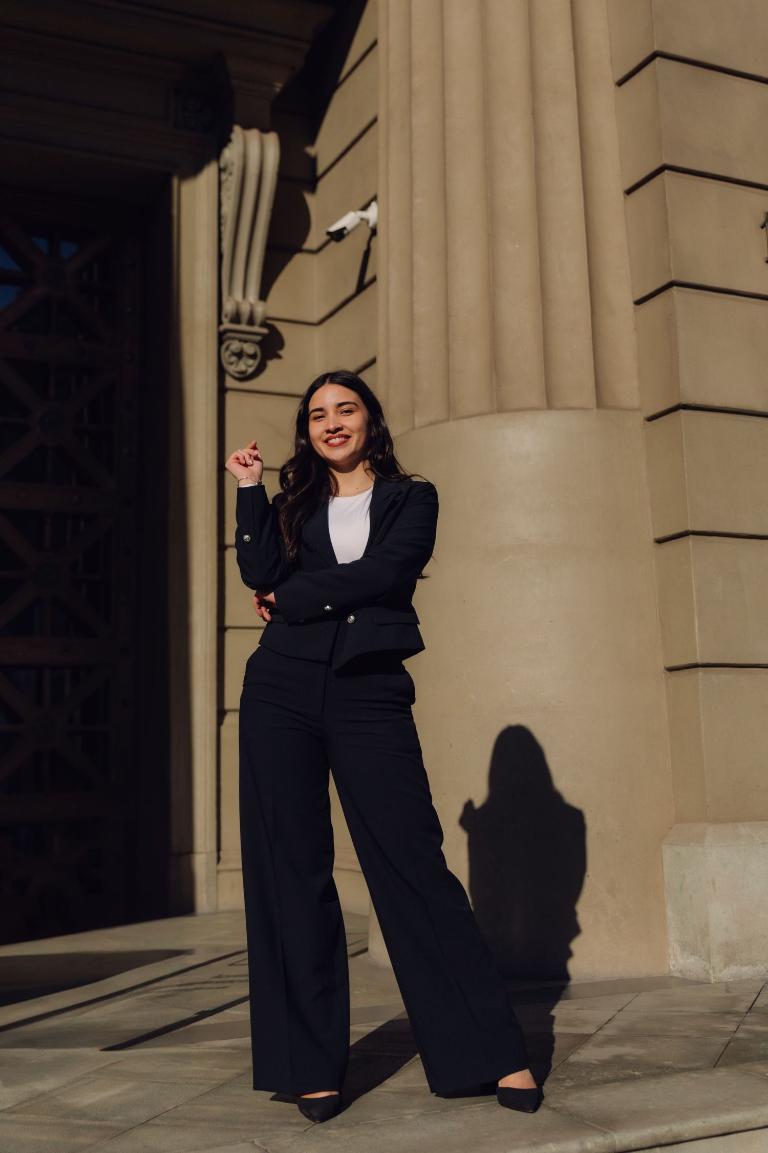 Confident young woman in black suit posing beside a stone column in sunlight.