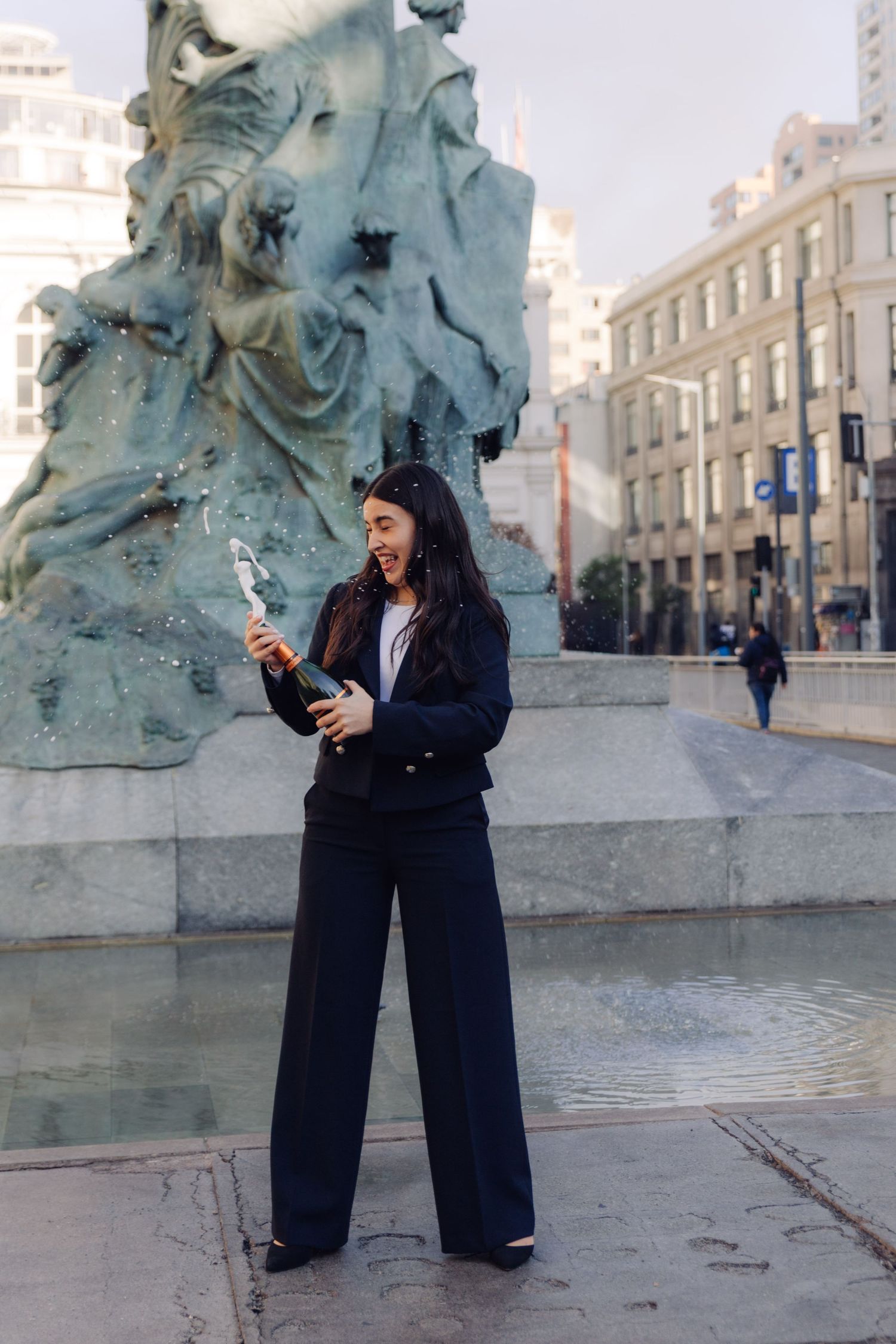 Young woman in black suit popping champagne bottle near an ornate city fountain.