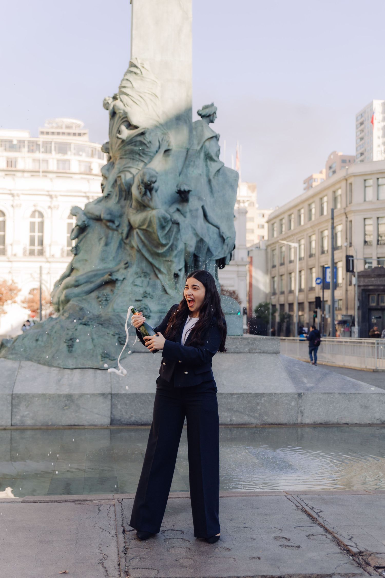 Excited woman in black blazer celebrating with champagne by a bronze city fountain.