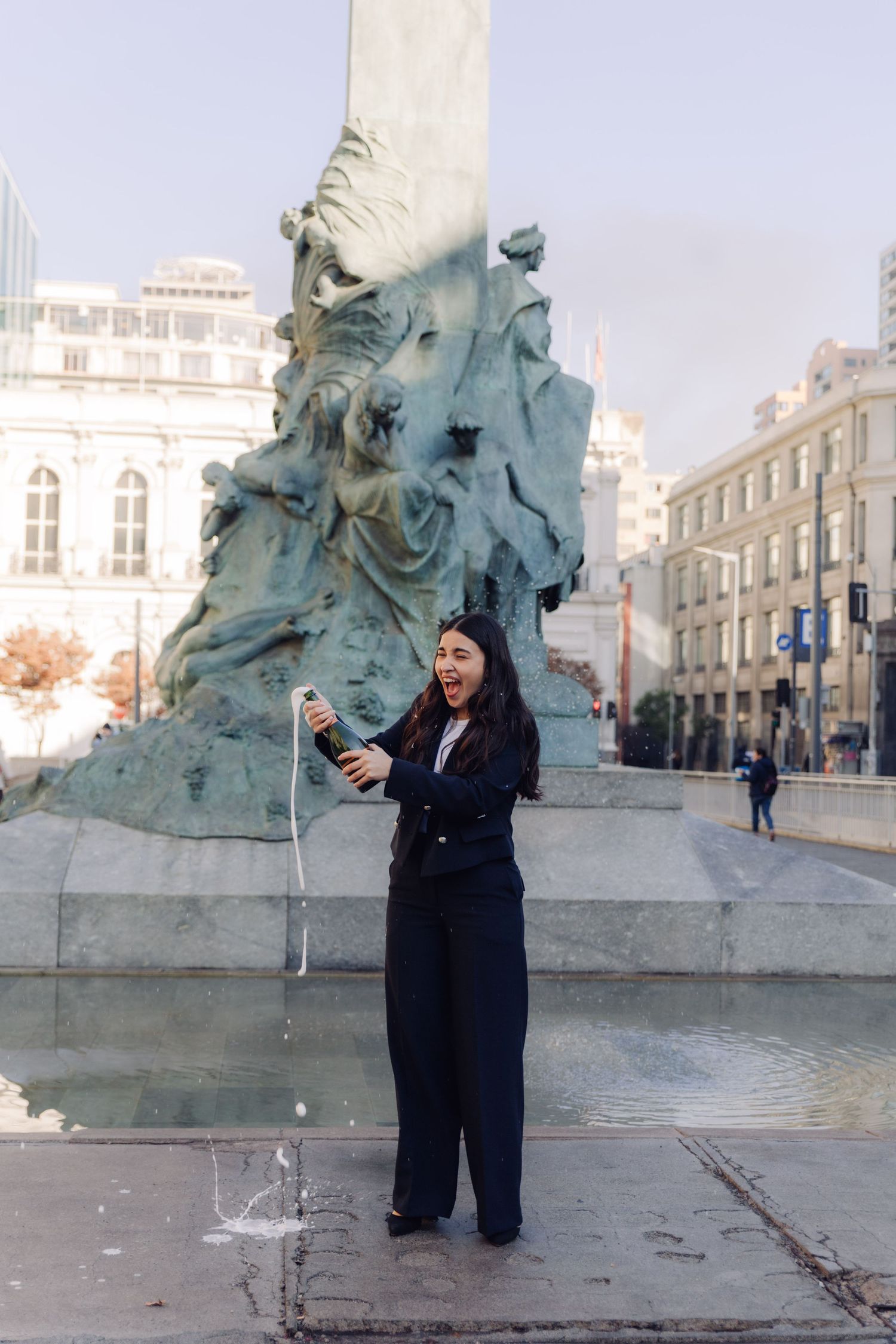 Joyful young woman in black suit spraying champagne beside an ornate bronze fountain.