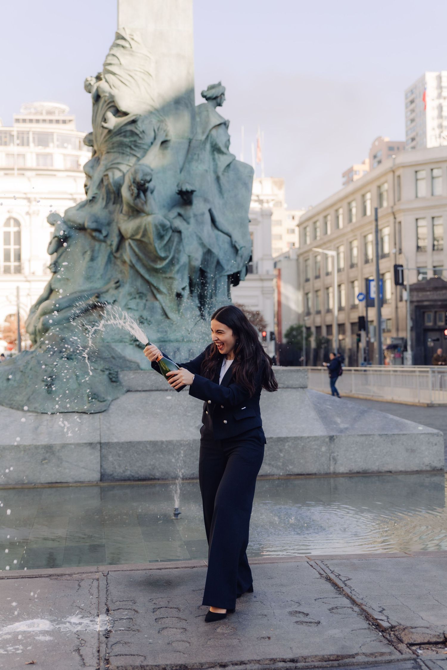 Woman in navy suit popping champagne bottle near bronze fountain statue in city square.