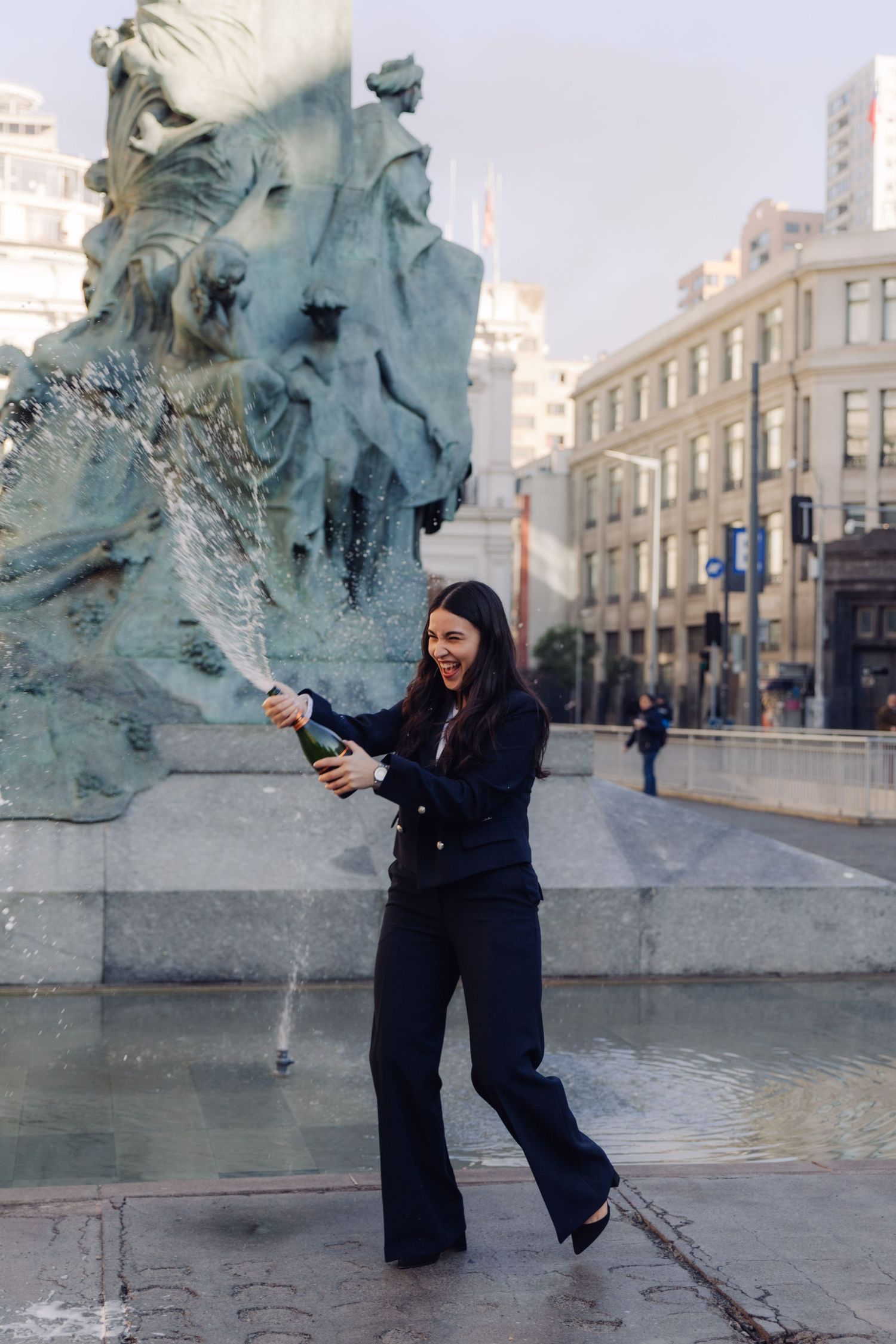 Excited woman spraying champagne from bottle beside ornate bronze monument outdoors.