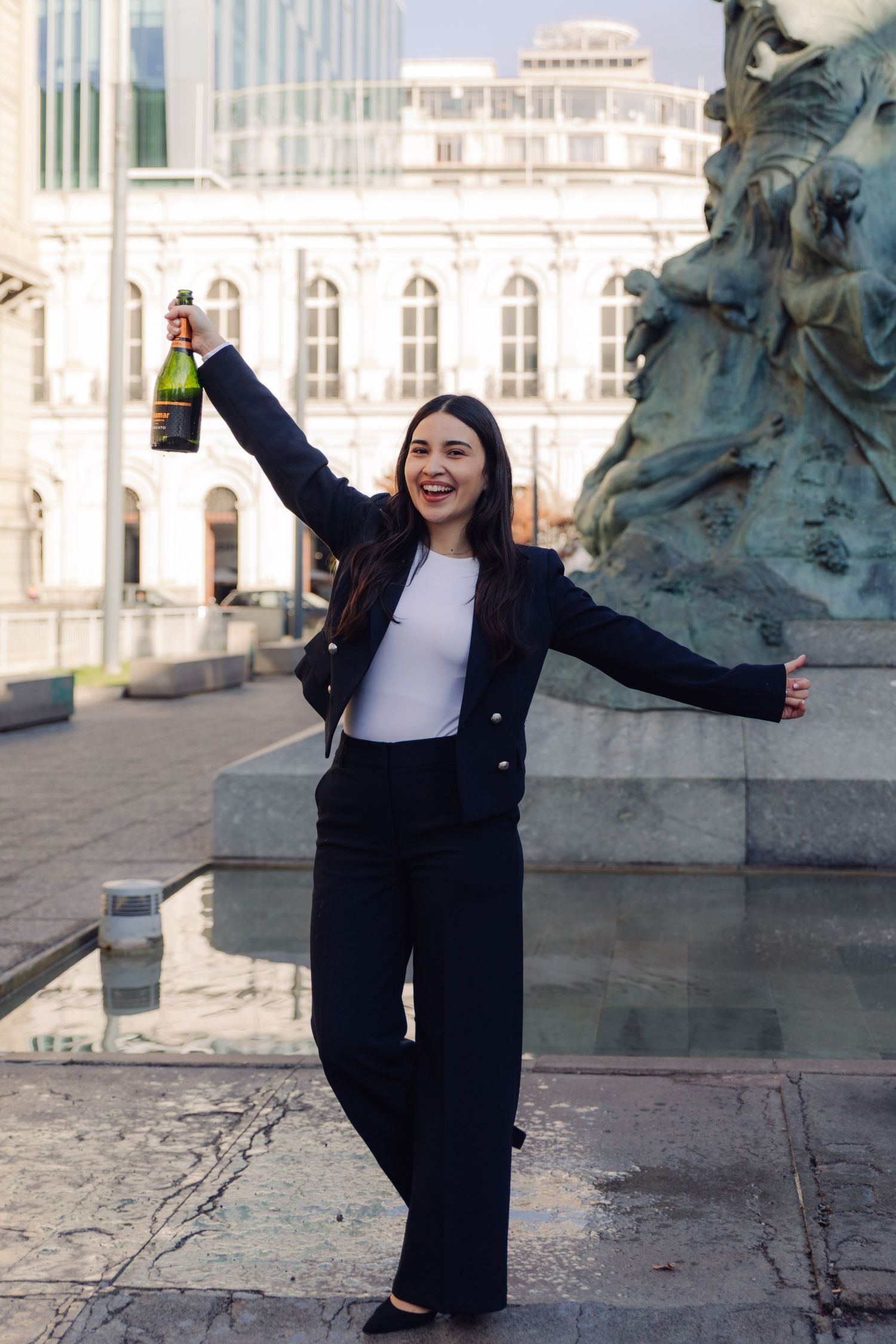 Smiling woman in navy blazer raising champagne bottle triumphantly near bronze fountain statue.