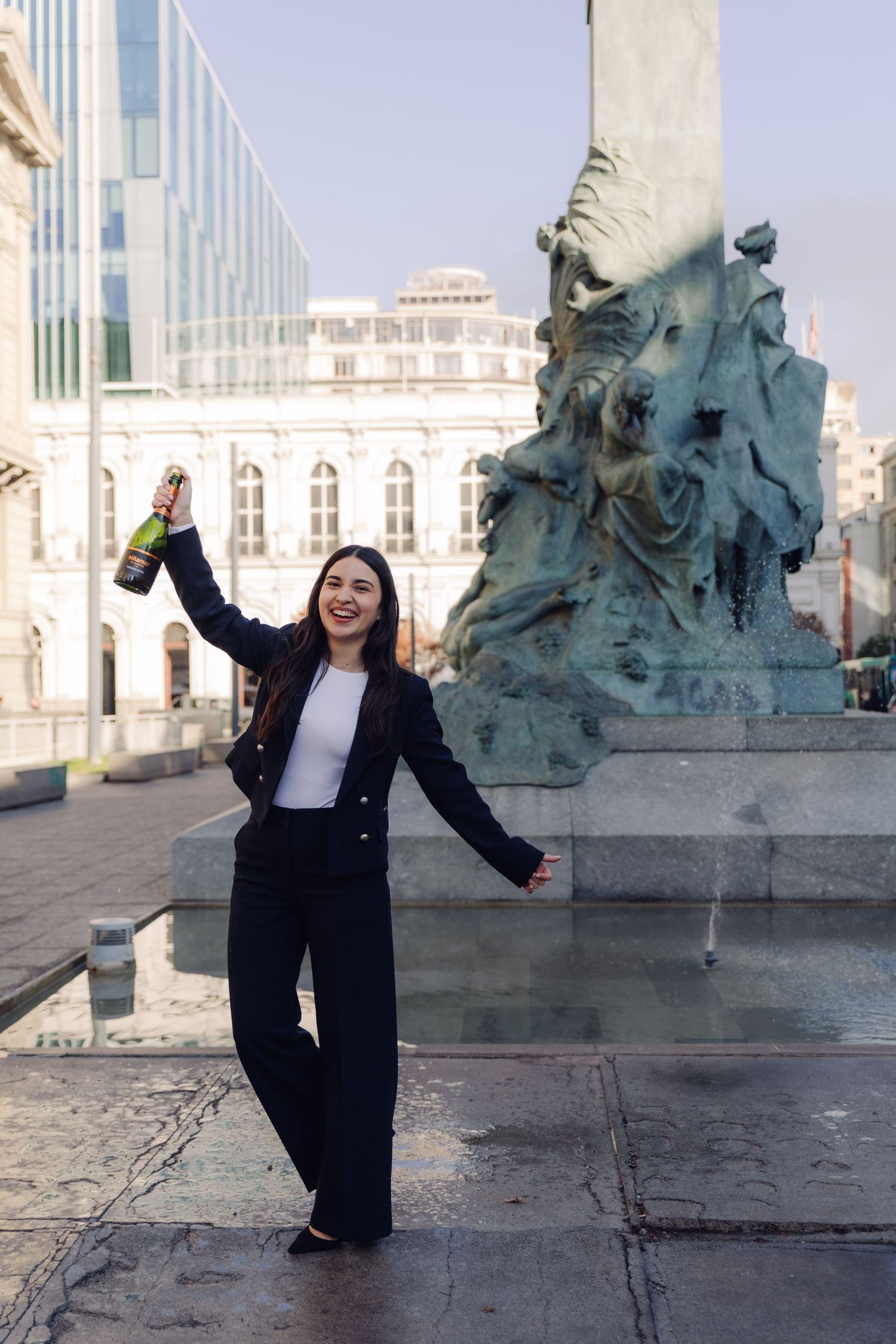 Happy woman celebrating with champagne bottle raised beside ornate bronze monument in city plaza.