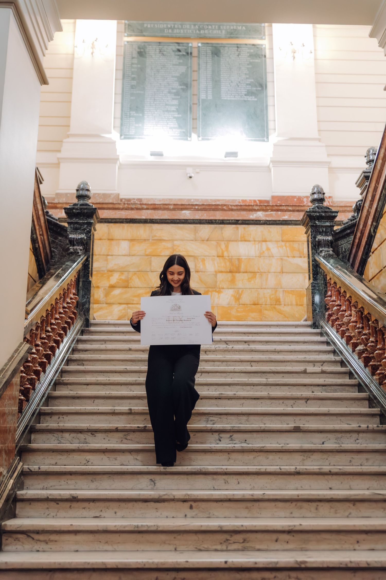Young woman descending grand marble staircase holding white certificate document indoors.