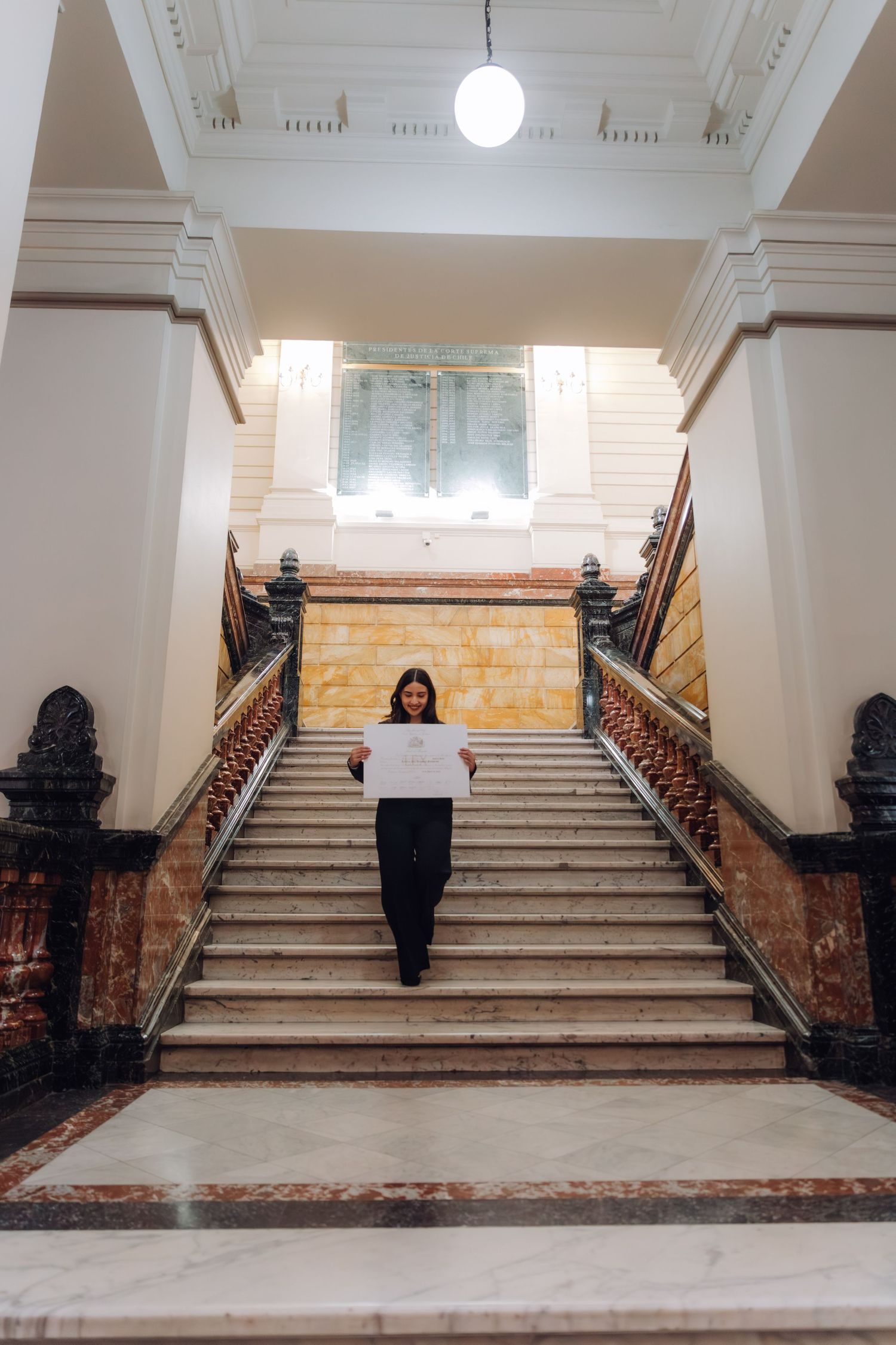 Woman standing on elegant marble staircase holding white document in classical building interior.