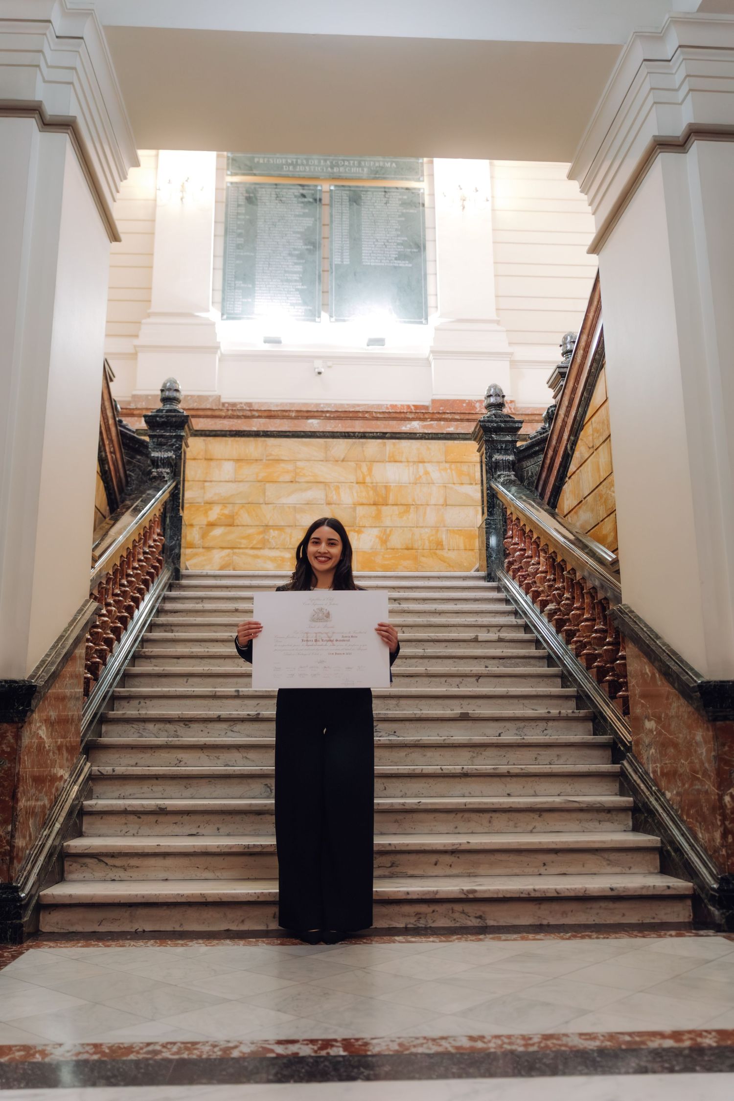 Smiling woman proudly displaying diploma certificate on grand marble staircase in historic building.