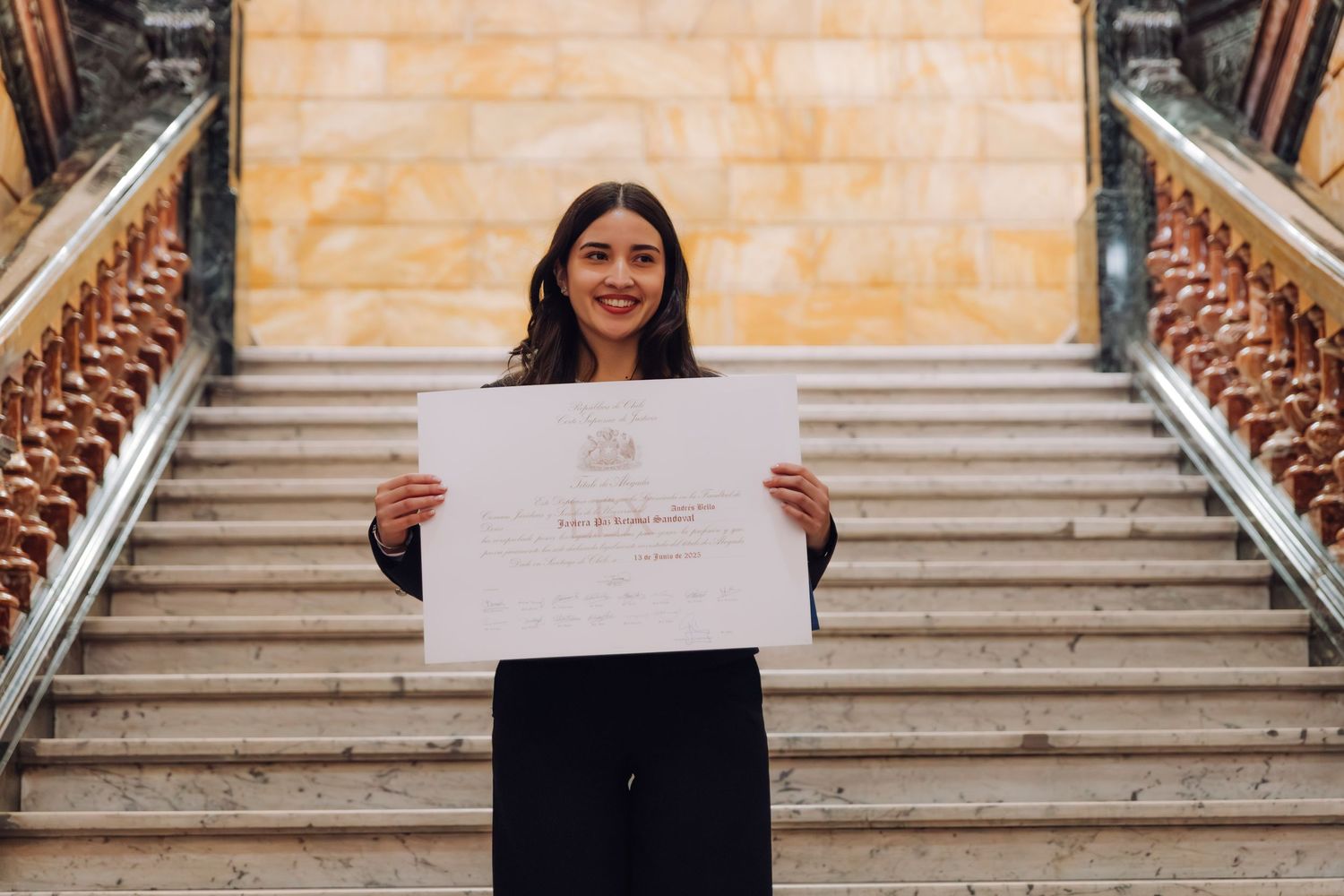 Young woman smiling and holding official diploma certificate on ornate marble staircase.