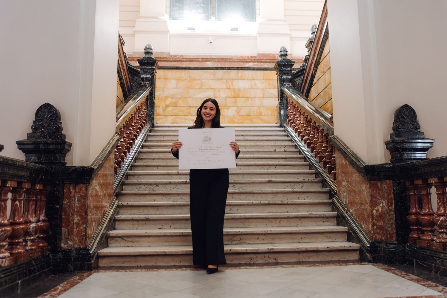 Woman standing before grand marble staircase holding white certificate document in formal building.