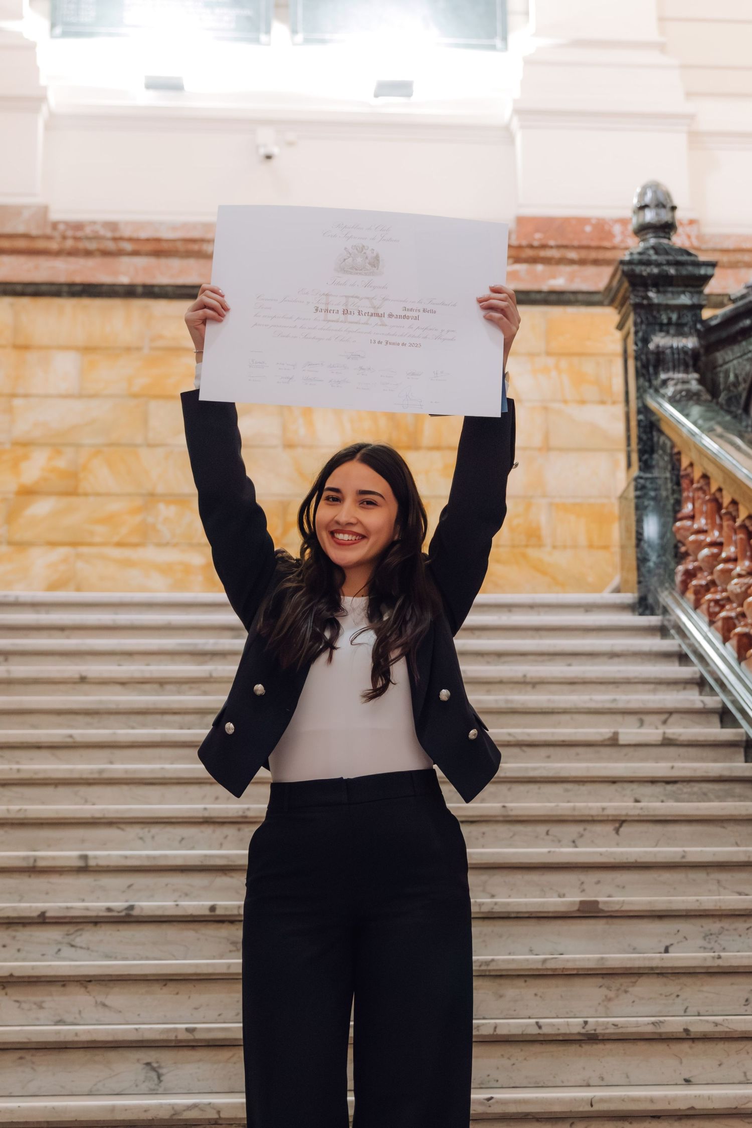 Jubilant woman raising diploma certificate overhead on marble staircase in elegant building.