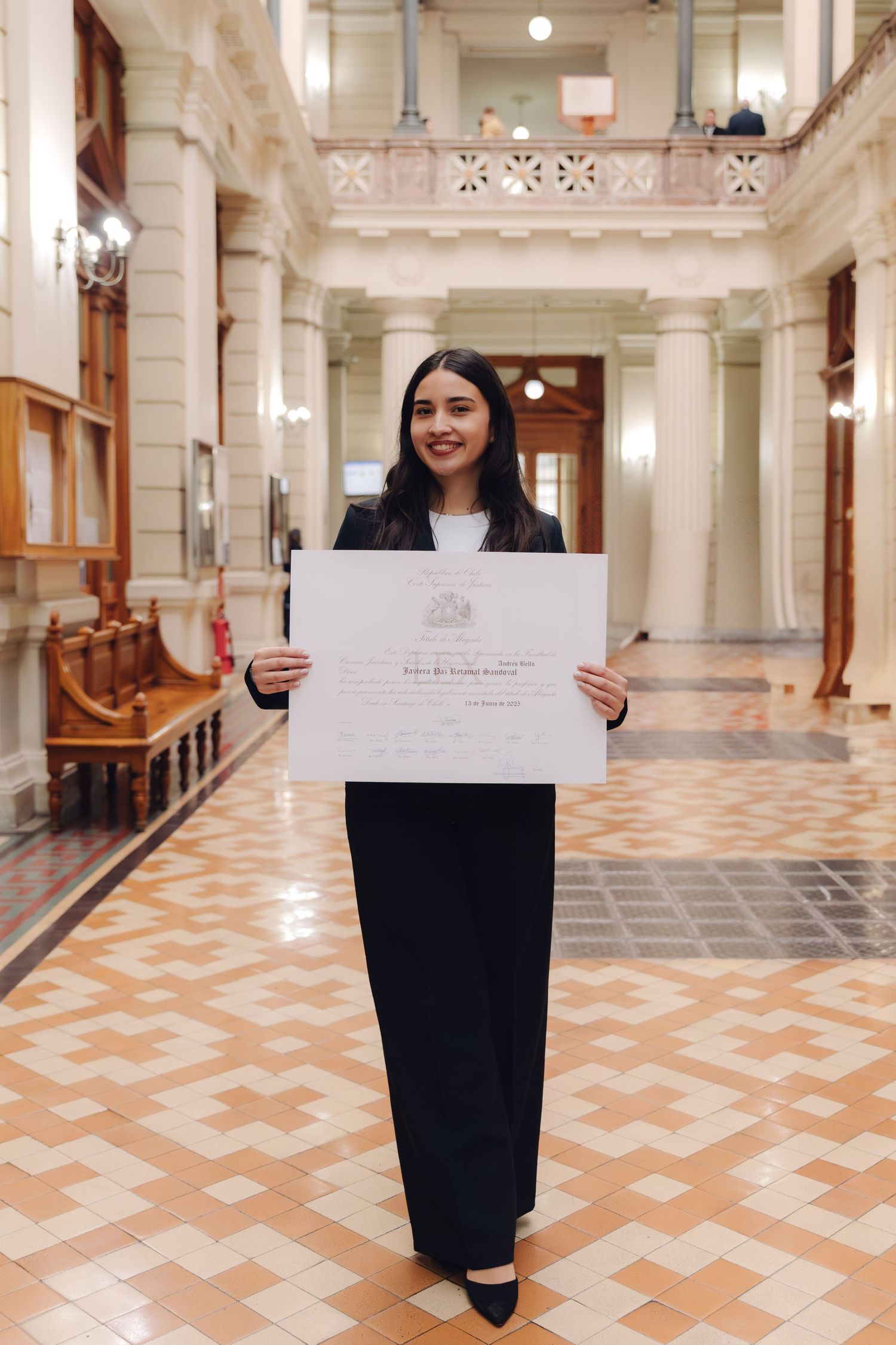 Smiling graduate holds diploma in ornate university hallway wearing black suit.