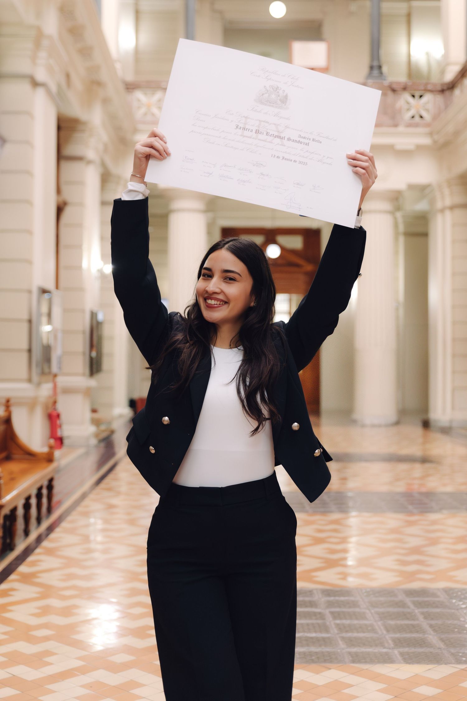 Young woman in black blazer joyfully raises diploma overhead in elegant hall.