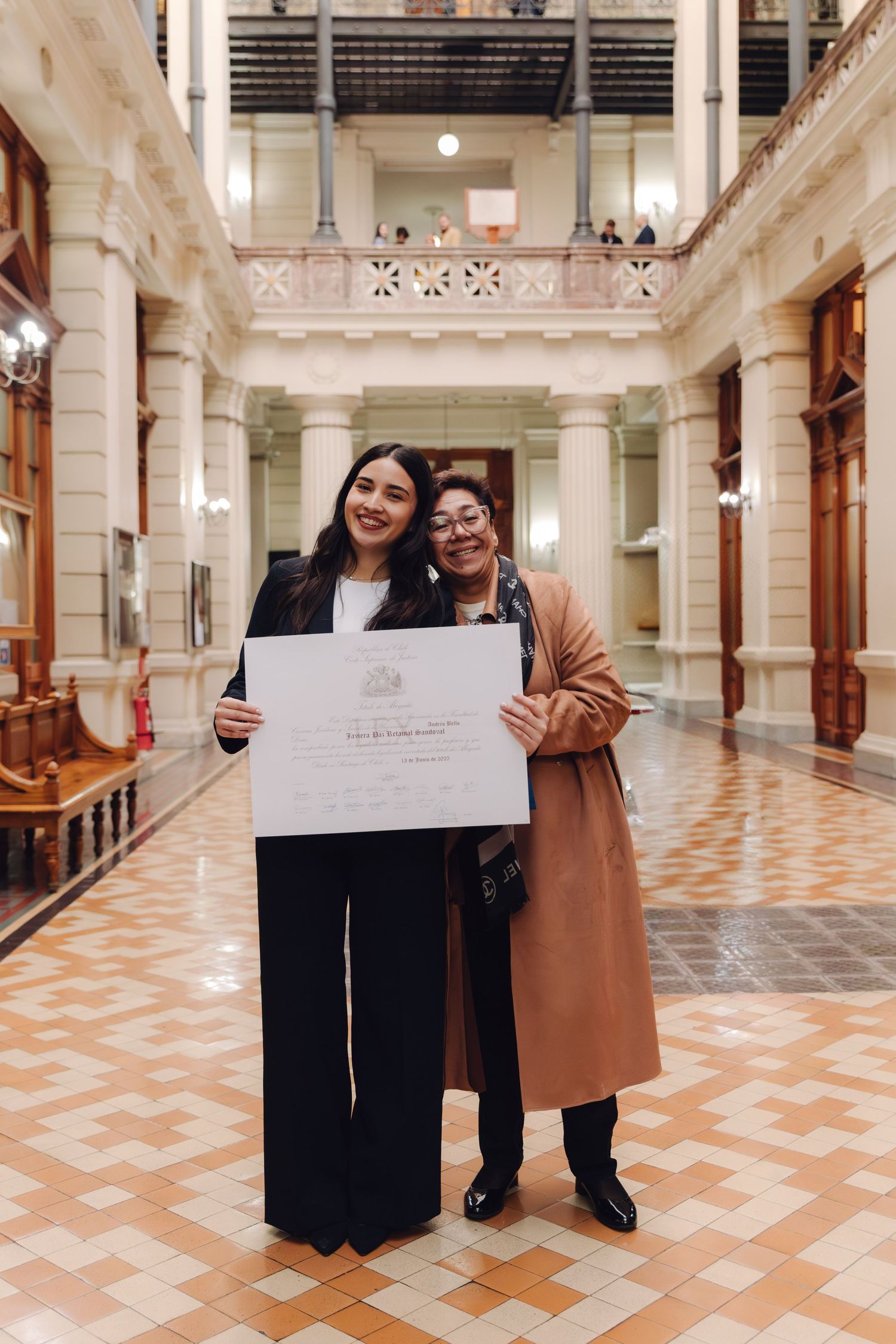 Two women smile holding graduation diploma in elegant tiled hallway.