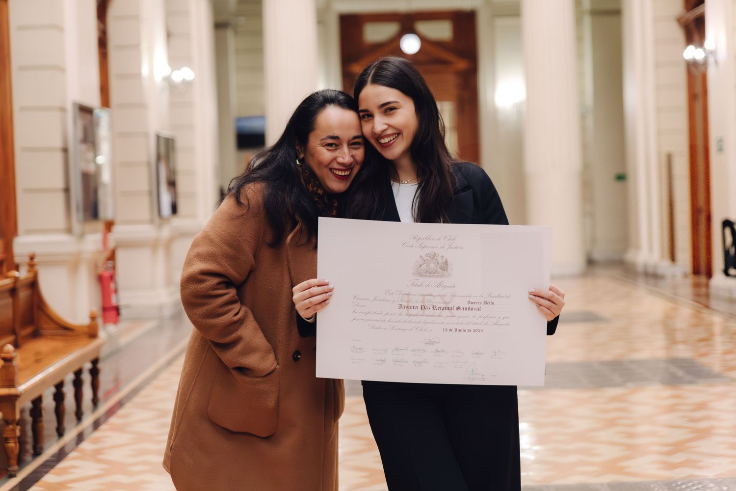 Two women laugh together holding graduation diploma in ornate hallway.