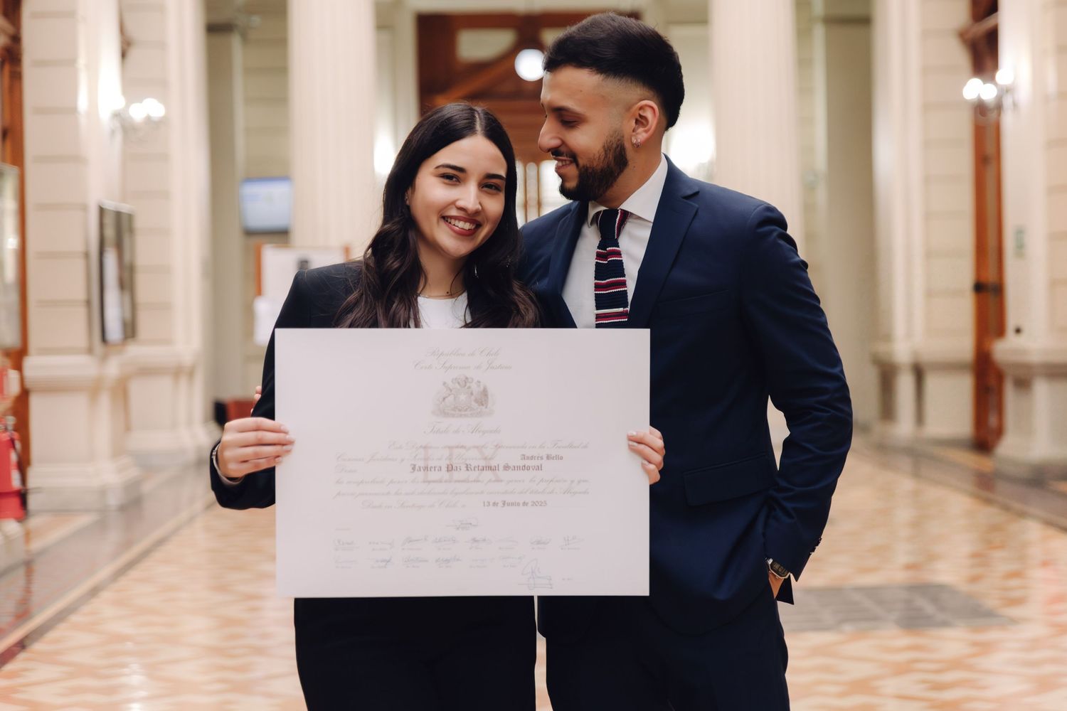 Couple in formal attire sharing a romantic moment while holding a graduation diploma certificate indoors.