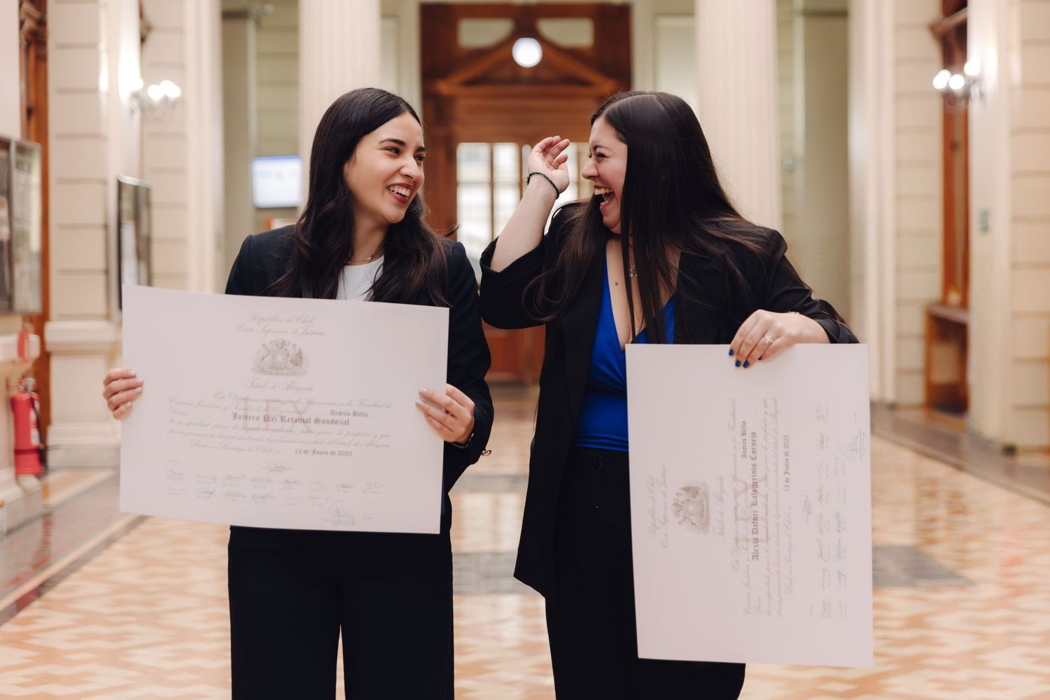 Two young women in black blazers laughing together while holding graduation diploma certificates in an elegant hallway.