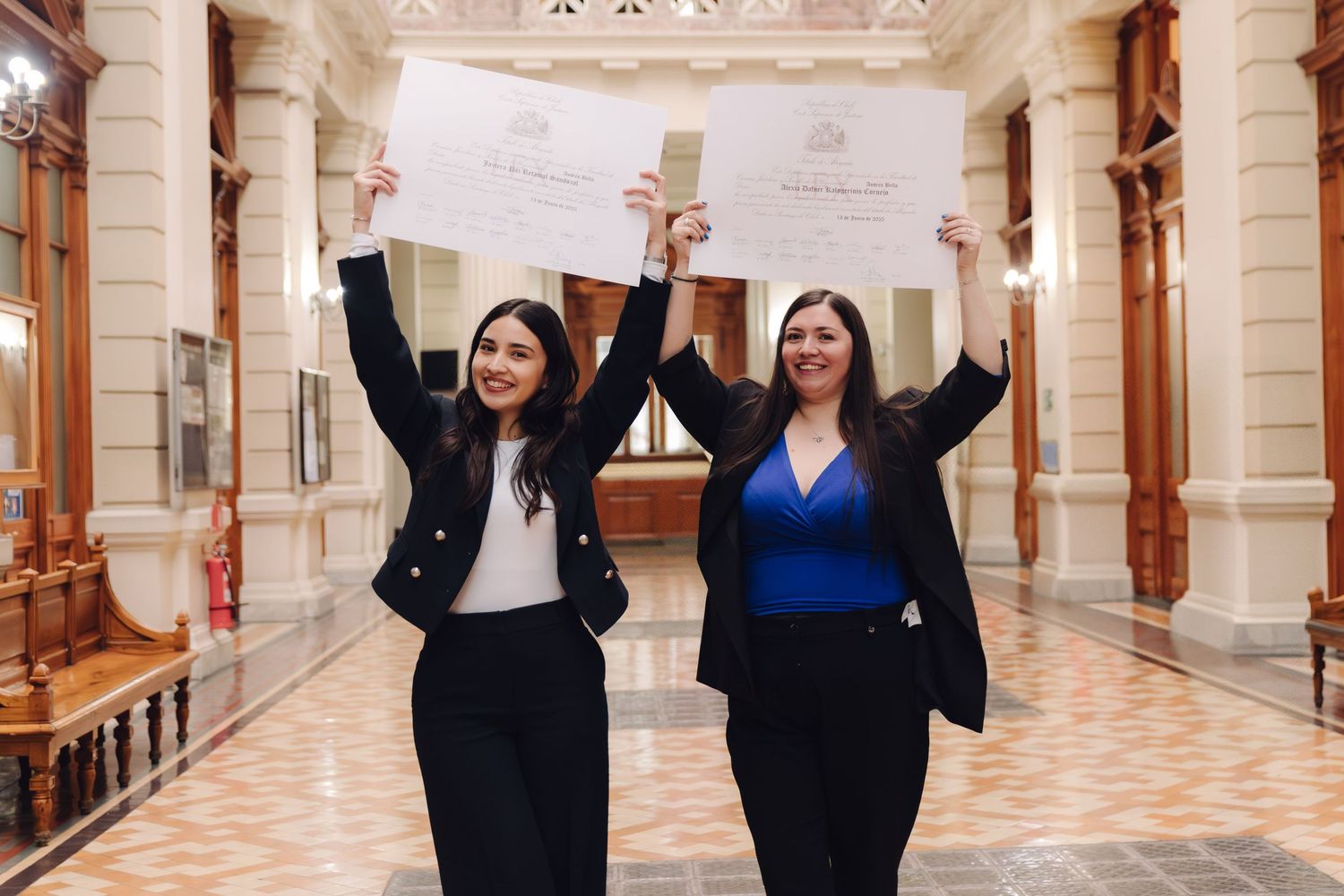 Two women celebrating graduation by raising their diploma certificates overhead in a grand interior hallway.