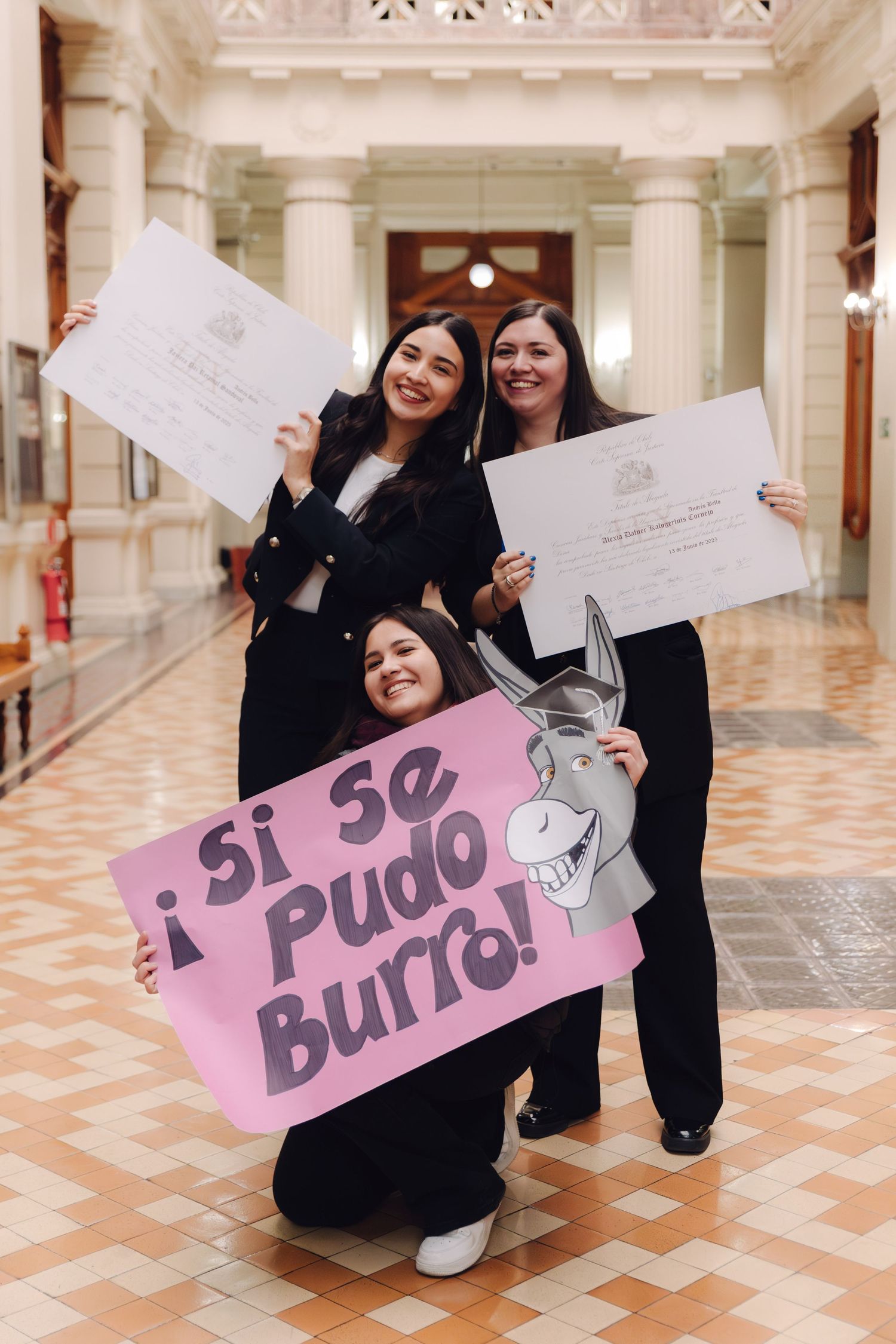 Three women celebrate graduation holding diplomas and a pink sign reading ¡Si se Pudo Burro! in an elegant hall.
