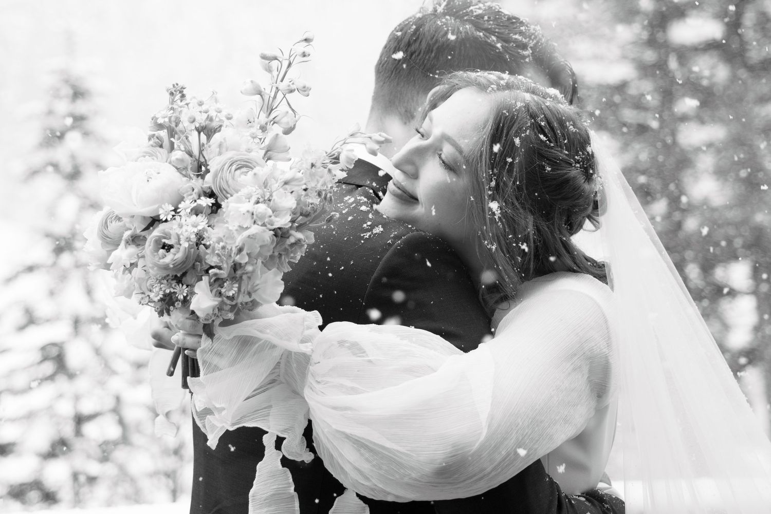 Black and white winter wedding photo of couple embracing in snowfall, bride holding bouquet wearing veil.