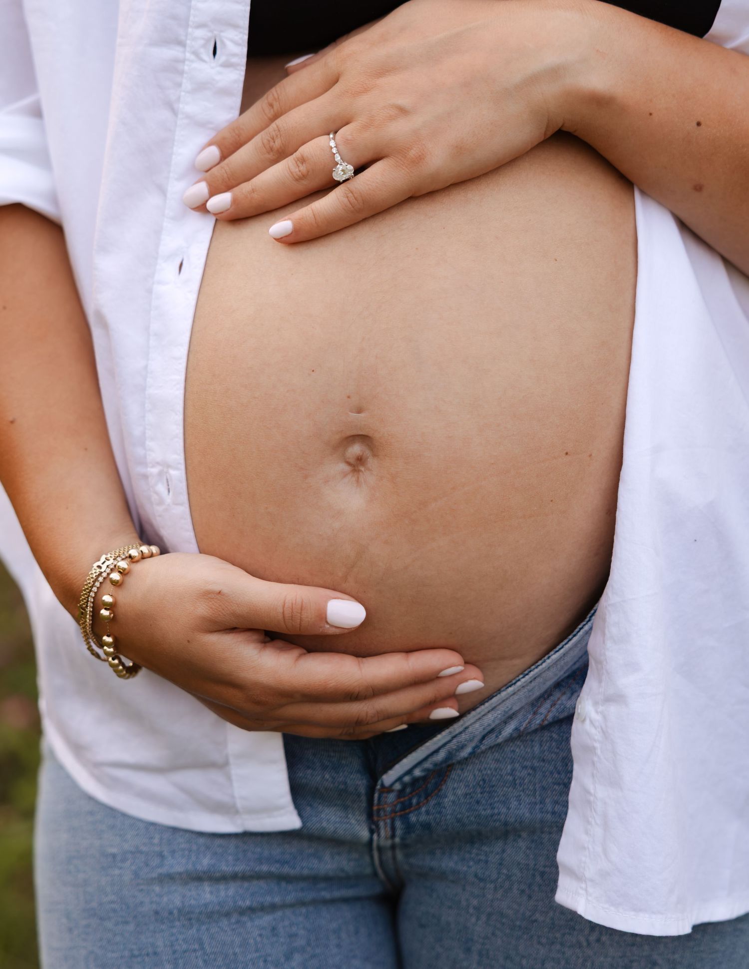 Pregnant woman in white shirt and jeans cradling baby bump, wearing engagement ring and gold bracelets, white manicure.