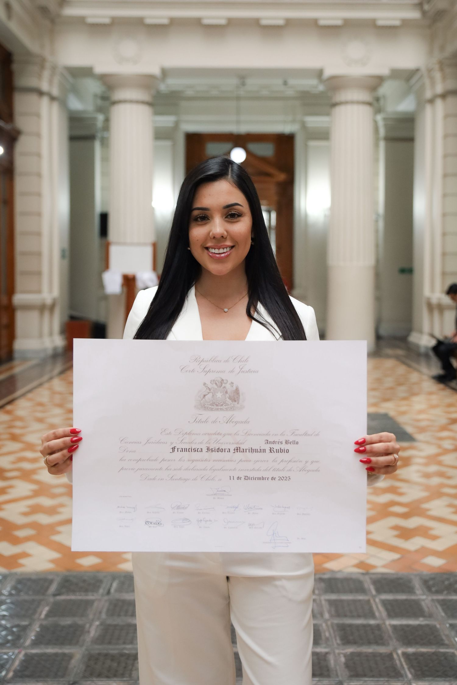 Smiling woman in white suit proudly holding official Chilean Supreme Court certificate indoors.